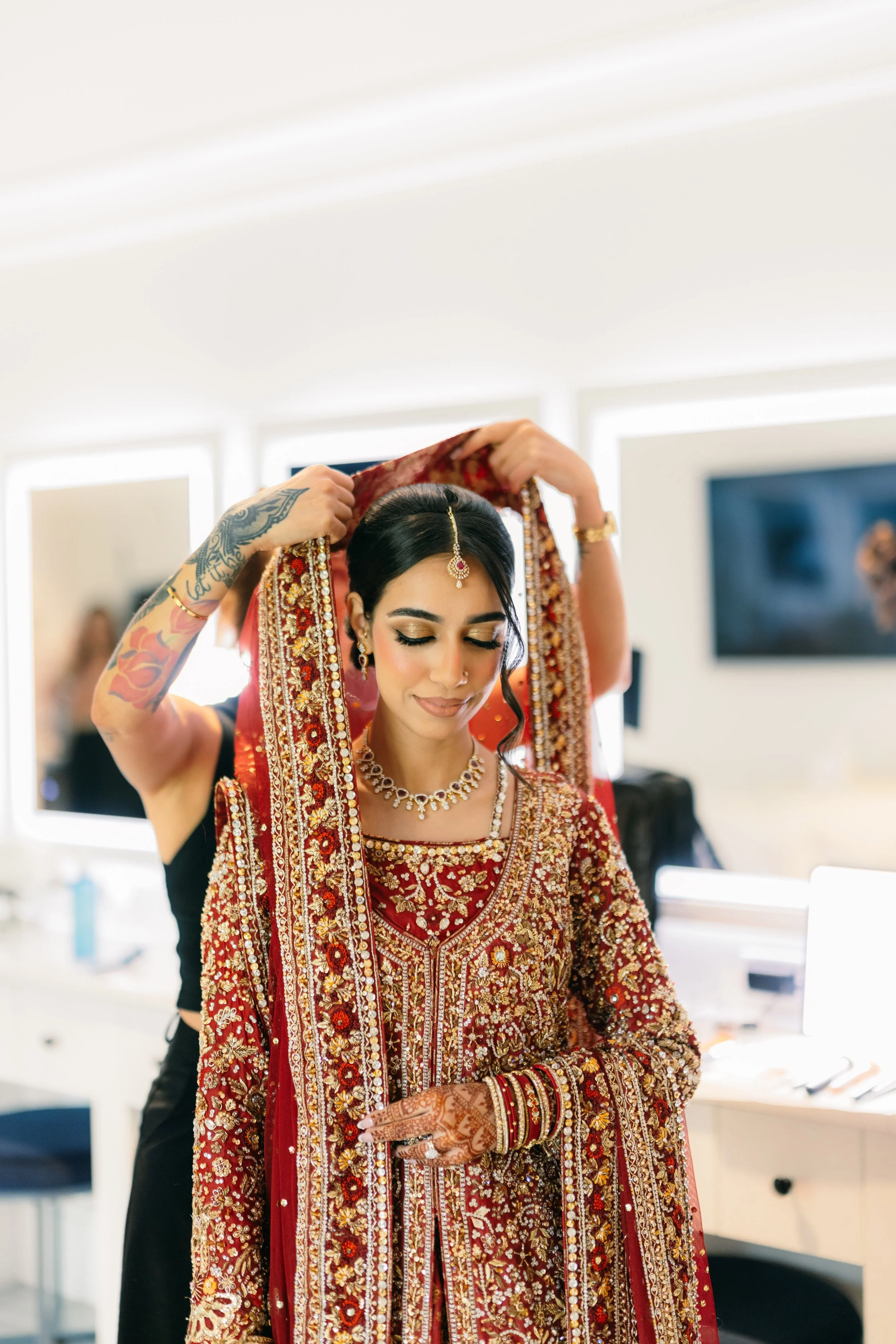 A bride in a traditional red and gold embroidered wedding dress, adjusting her headscarf with the help of another person in a well-lit room.