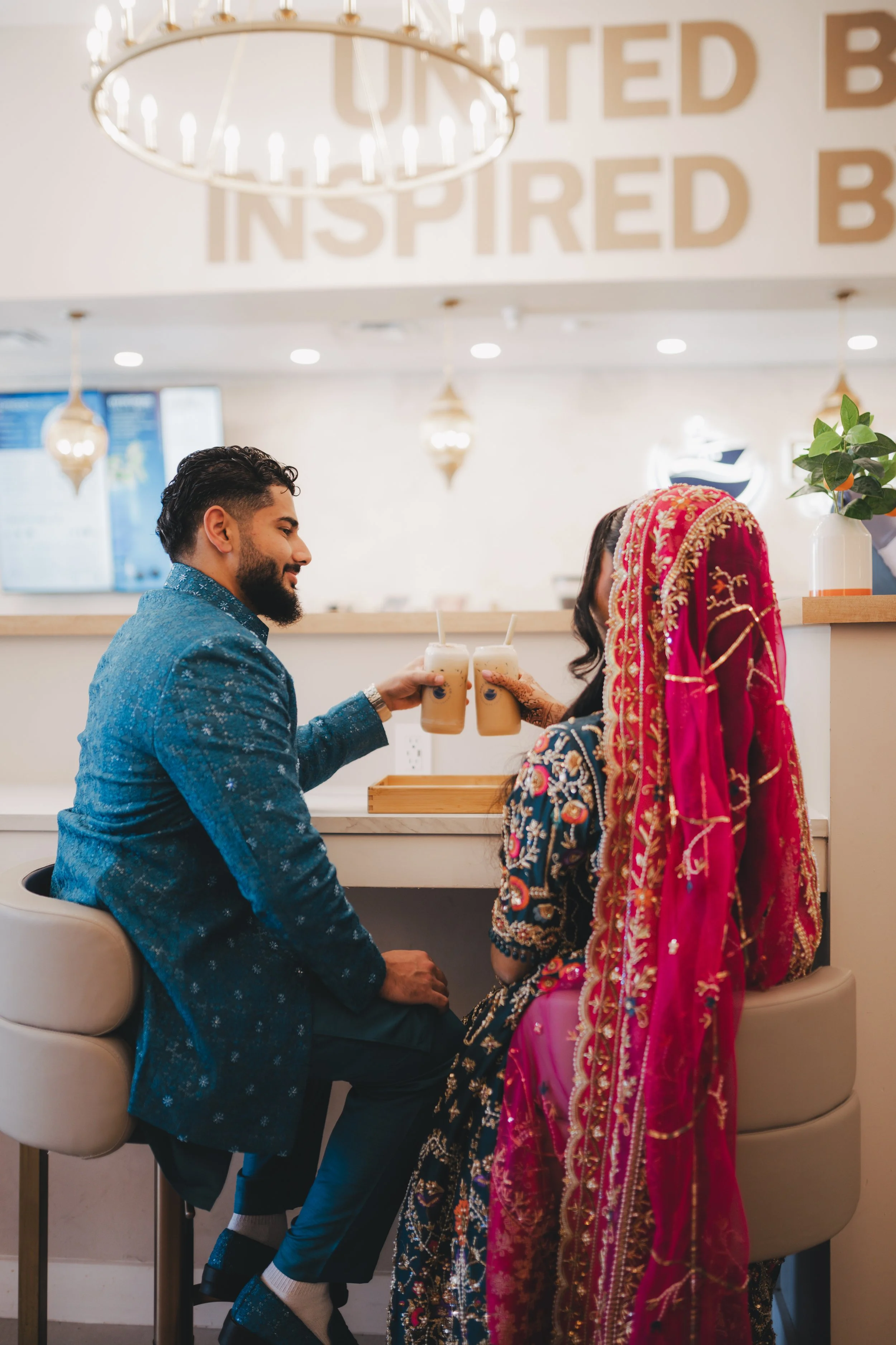 A man and woman dressed in traditional Indian attire sharing drinks at a cafe counter. The woman is wearing a vibrant pink and gold sari with a matching head covering, and the man is in a blue patterned blazer. They are smiling and clinking their dri