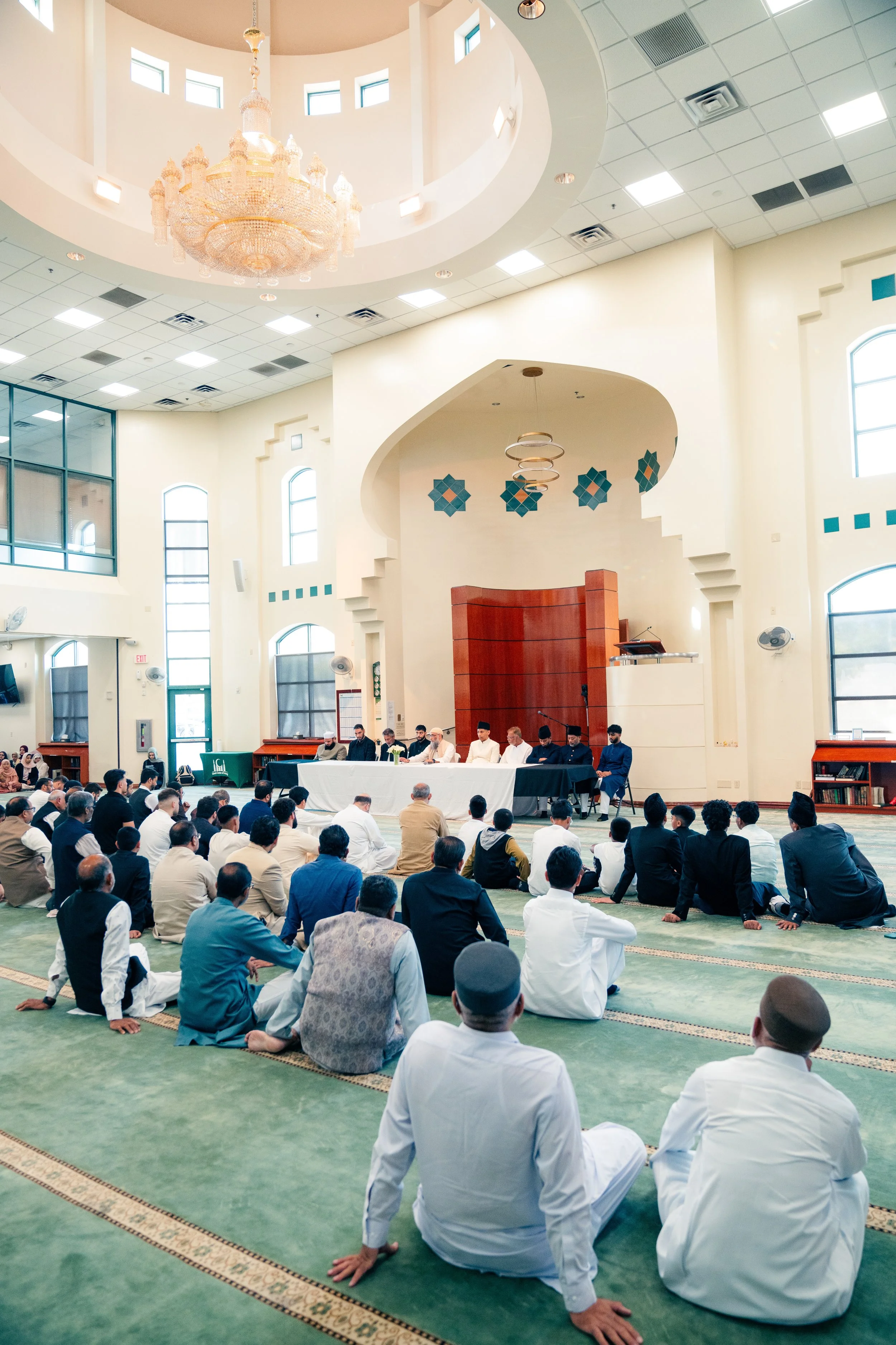 People gathered inside a mosque for a religious service, with some sitting on the green prayer carpet and others attending from a raised platform.
