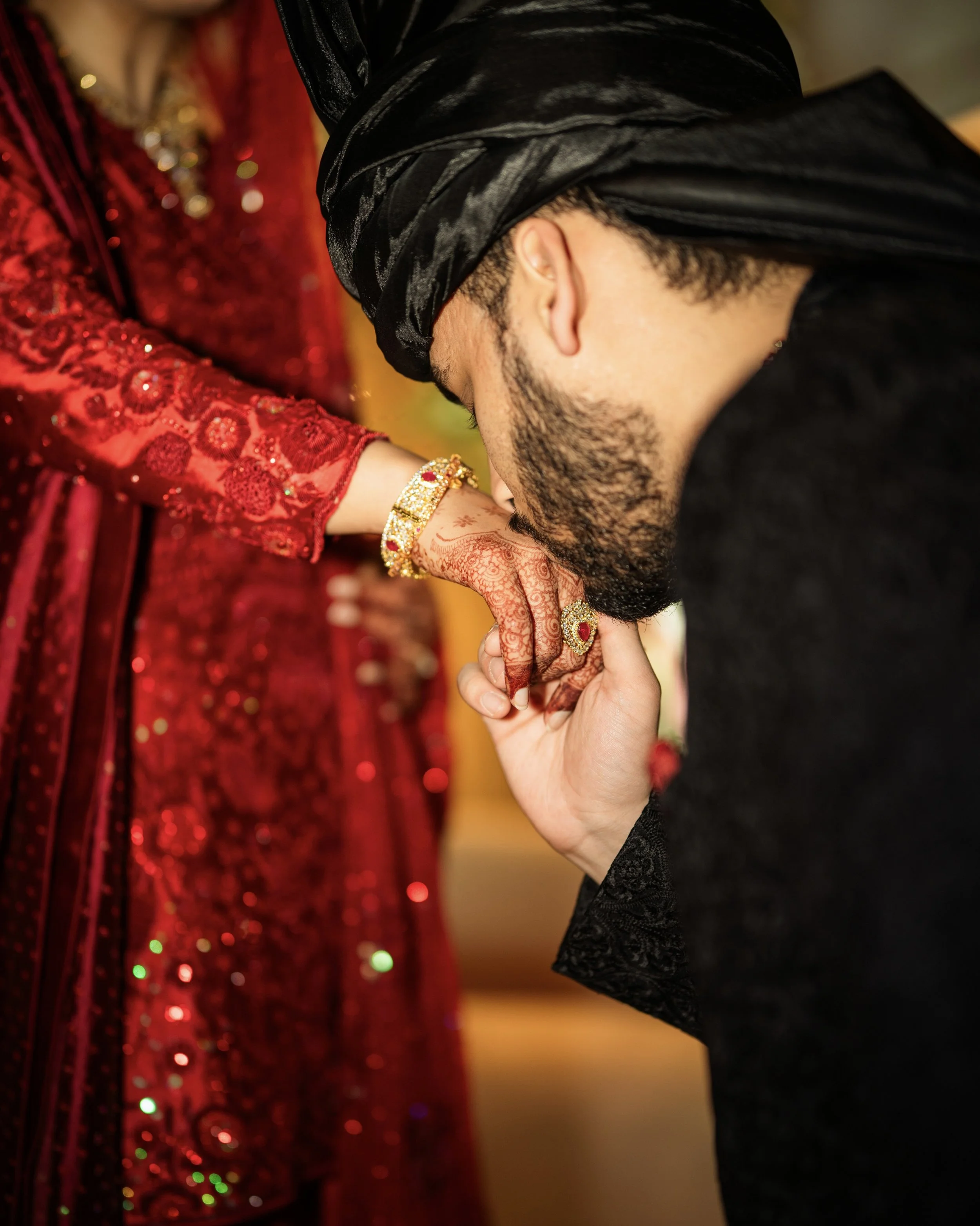 A man in traditional attire is kissing the hand of a woman dressed in a red embroidered outfit, adorned with jewelry and henna designs.