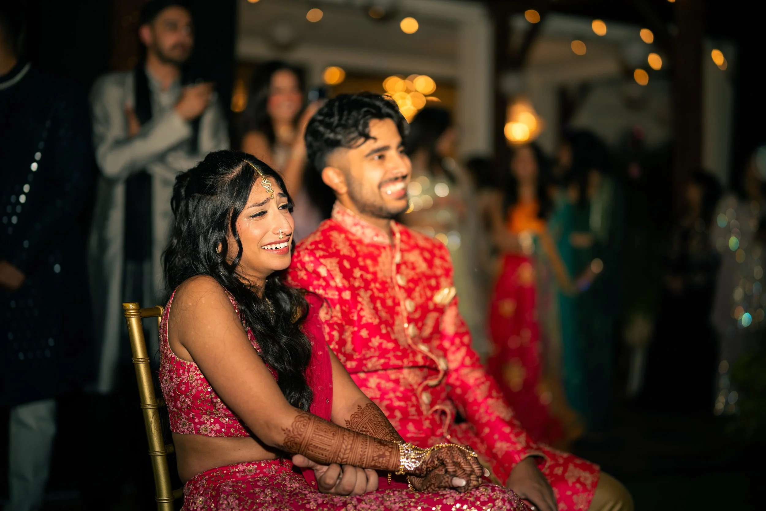 A joyful Indian wedding reception with a woman and man in traditional attire sitting together, surrounded by guests.