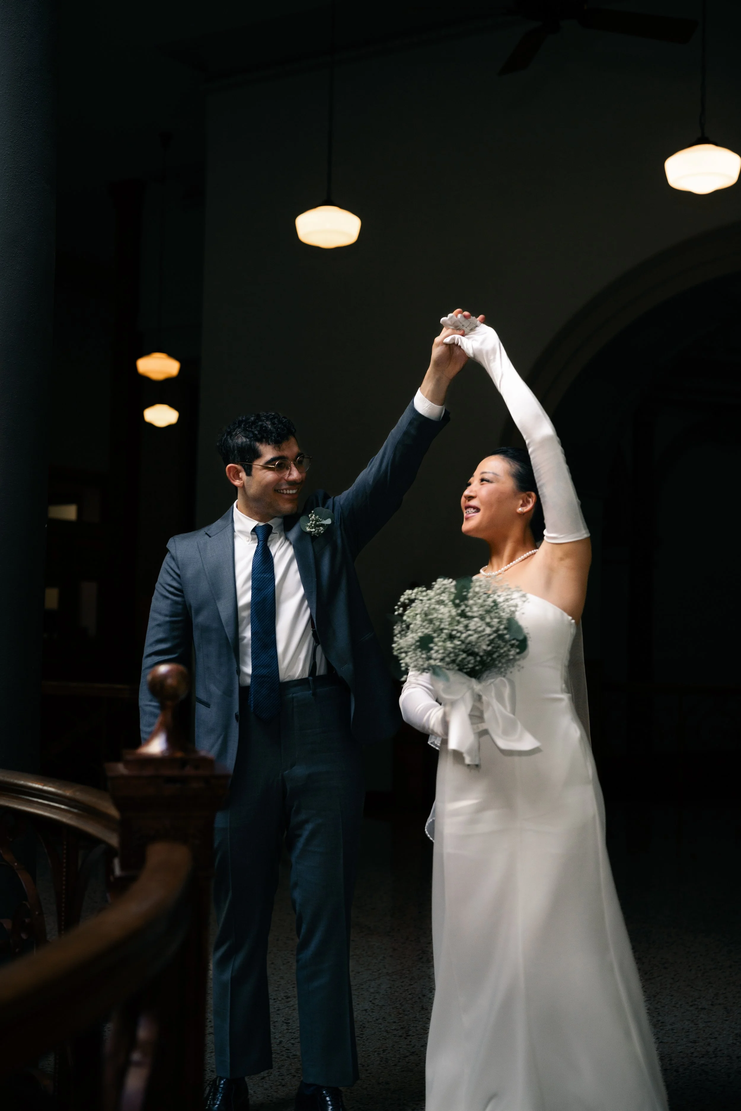 A bride and groom dancing together indoors, with the groom holding the bride's hand up while she holds a bouquet of flowers, both smiling. The lighting is warm and dim.