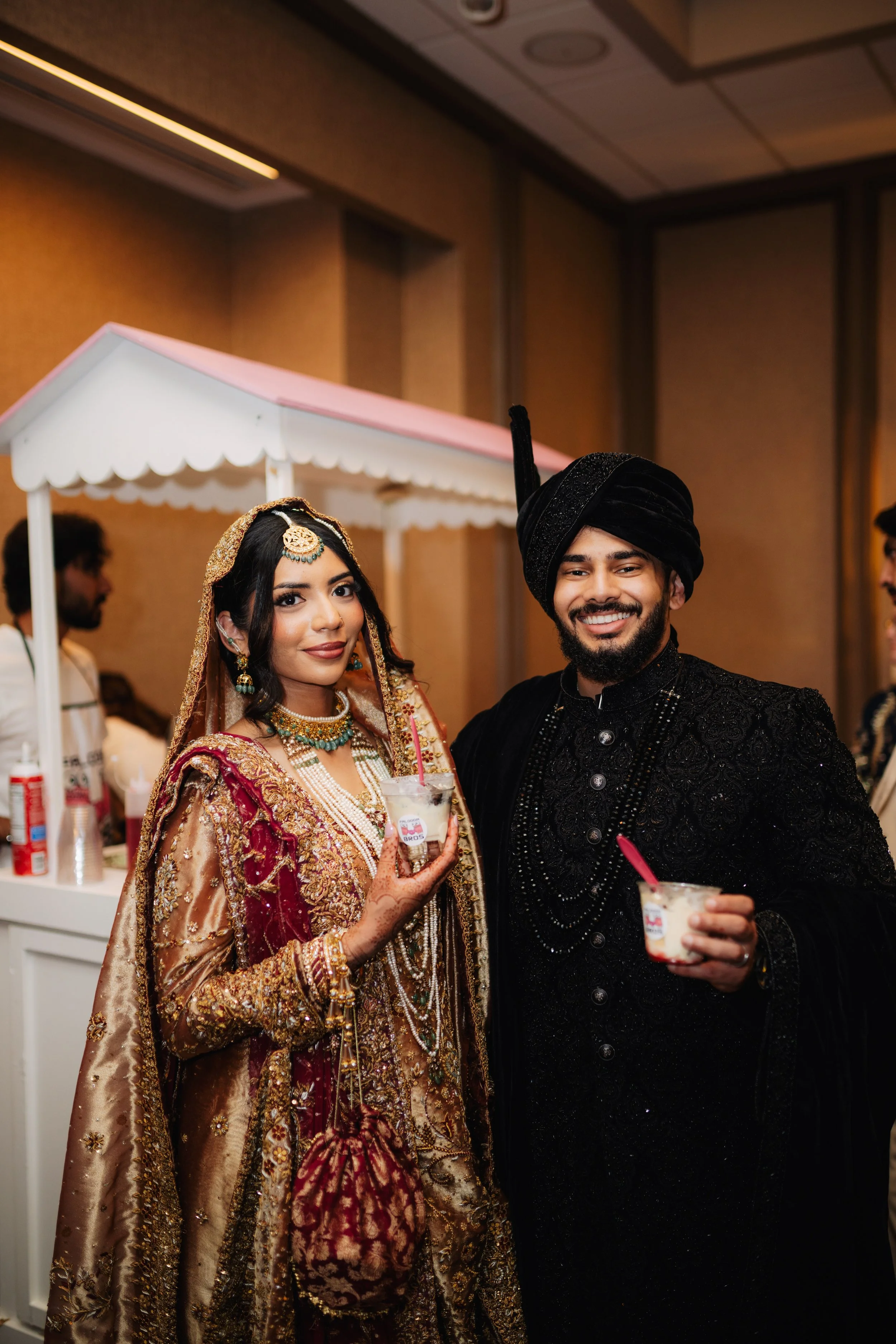 A couple dressed in traditional Indian wedding attire, smiling and holding drinks, standing in a decorated indoor venue.