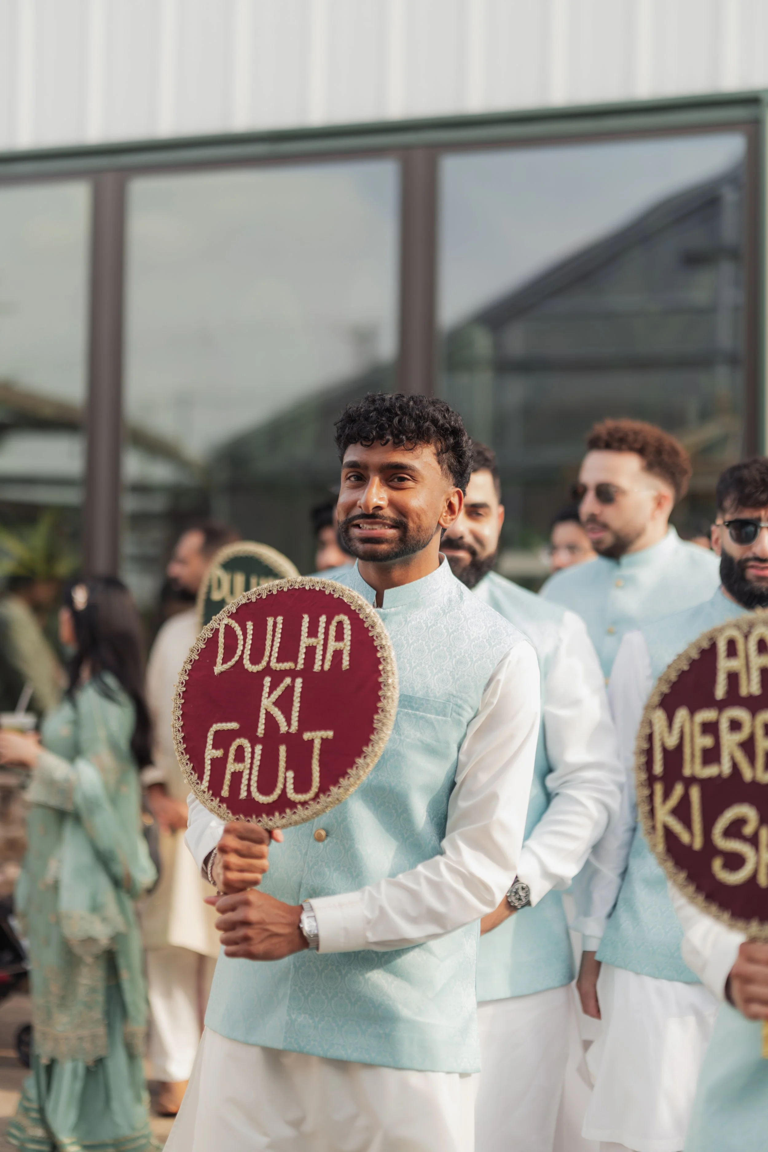 Man wearing traditional white and light blue outfit holding a circular sign with red background and gold border that reads 'DULHA KI FAULT' in an outdoor event with other similarly dressed men and women in the background.