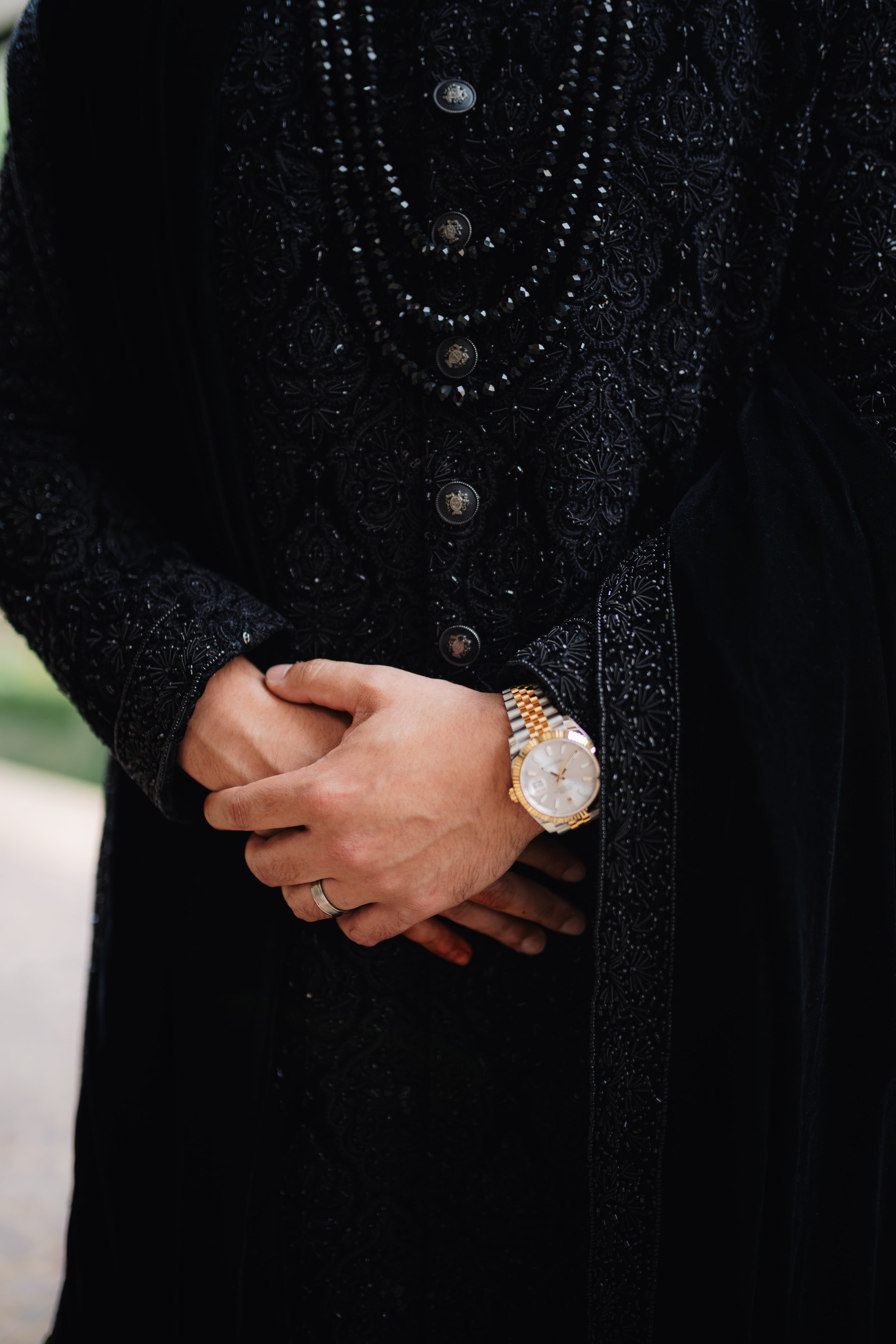Close-up of a person in a black embroidered jacket with jewelry and a gold wristwatch, standing outdoors with arms crossed.