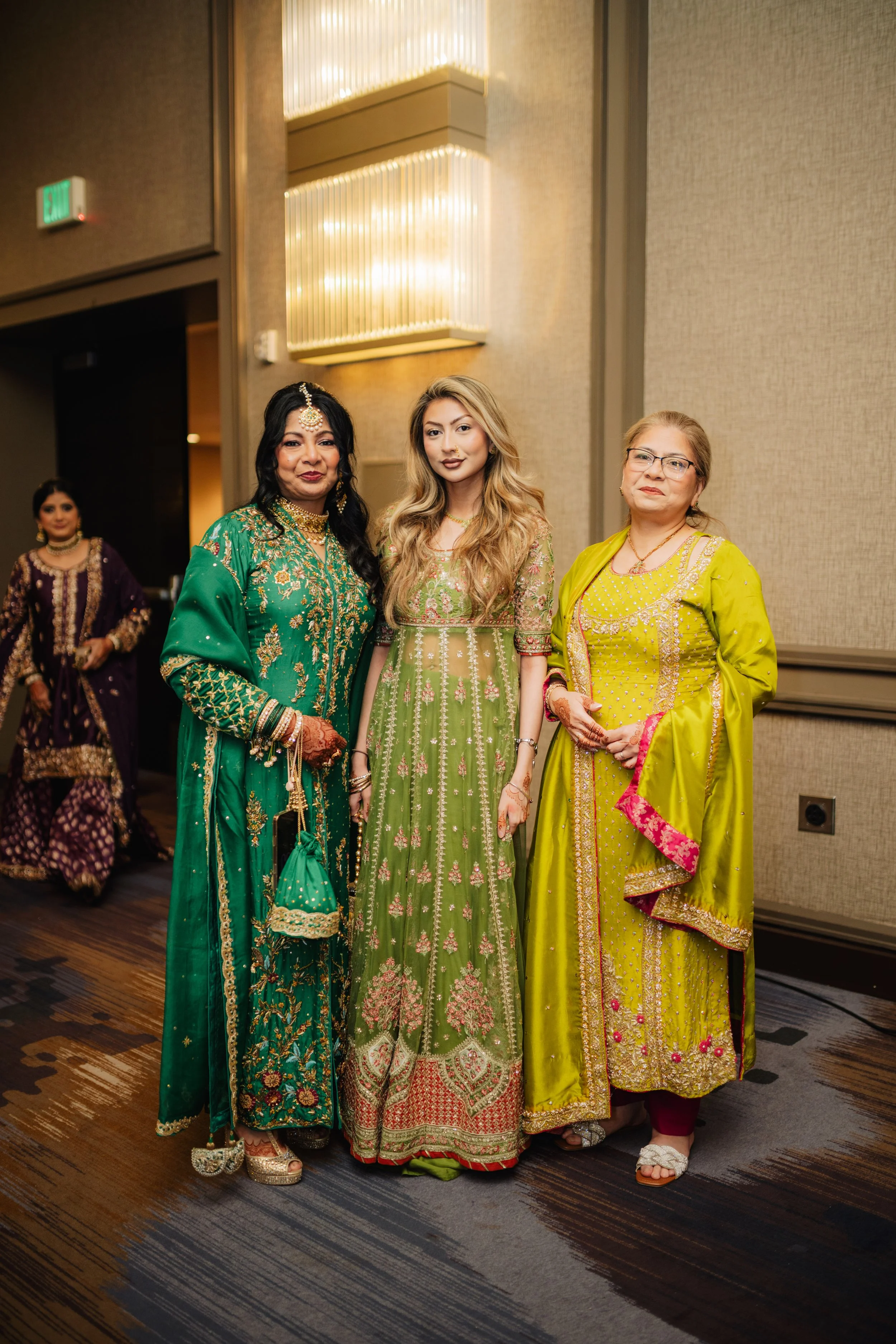 Four women dressed in traditional South Asian attire standing together indoors at a social event with beige walls and warm lighting.
