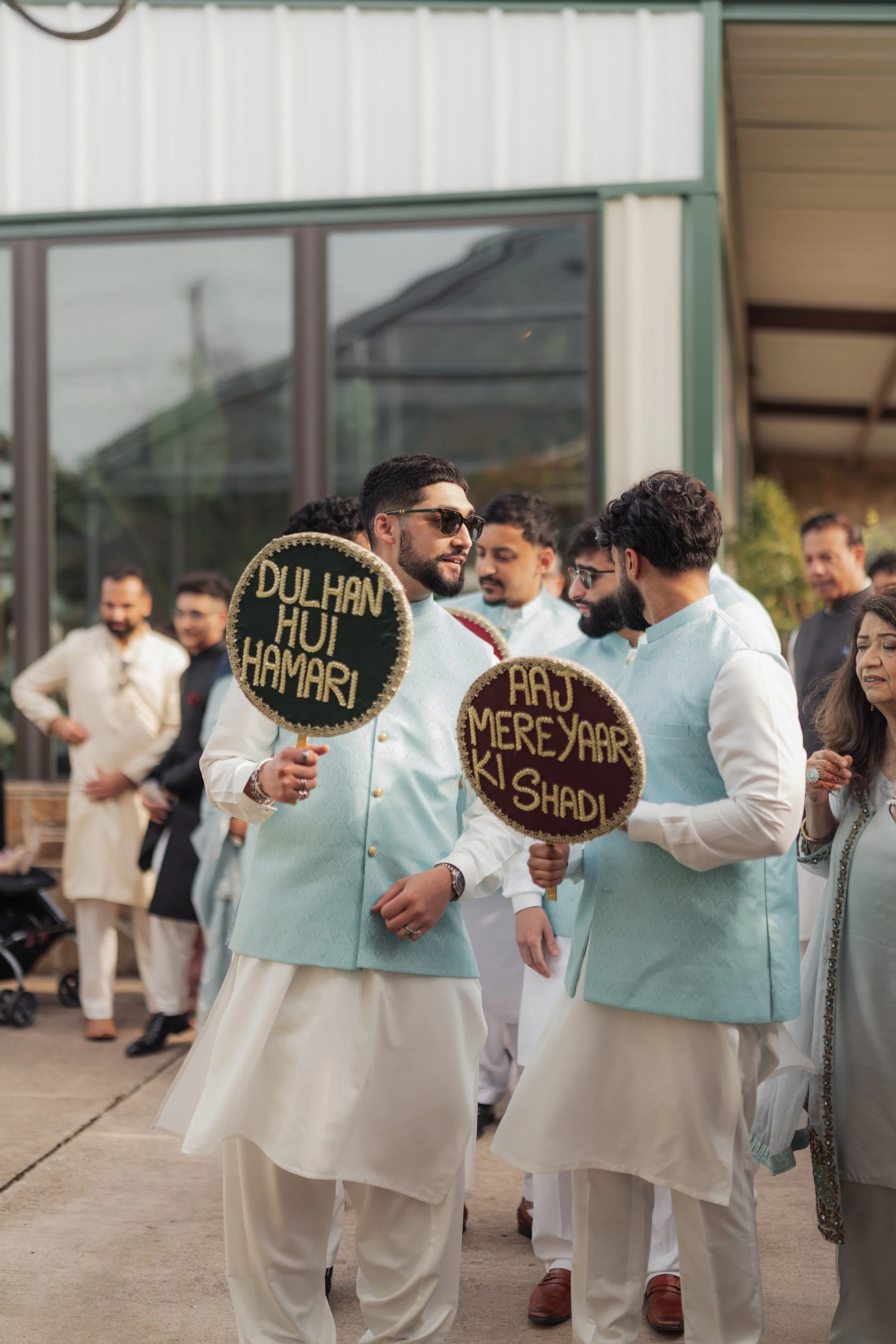 Group of men dressed in traditional Indian clothing participating in a cultural event or parade. Some holding decorative circular signs with Hindi-inspired text.