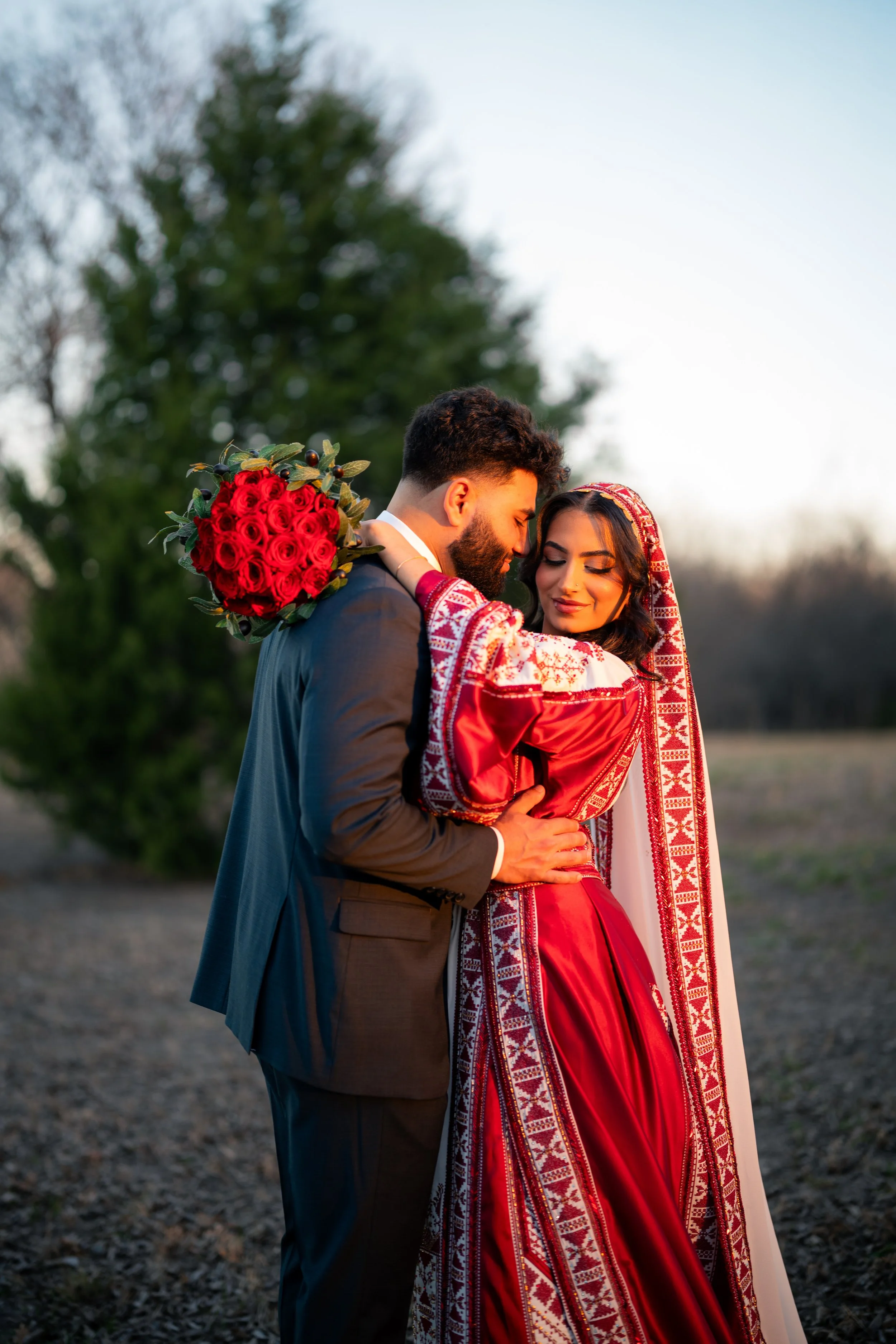 A couple embracing outdoors during sunset, the woman wearing a red traditional dress with embroidered details and a headscarf, and the man in a suit holding a bouquet of red roses.