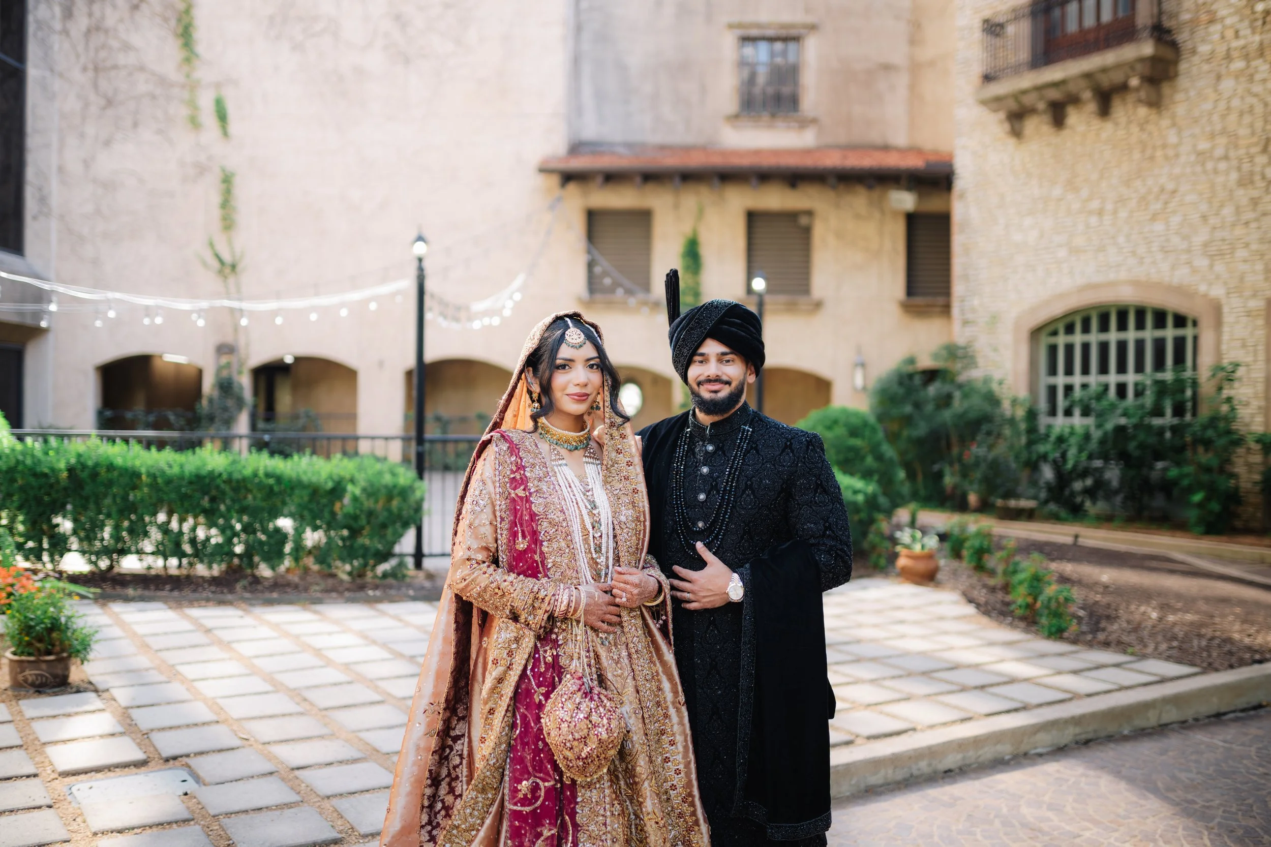A couple dressed in traditional Indian wedding attire standing outdoors in a courtyard with stone buildings and string lights.