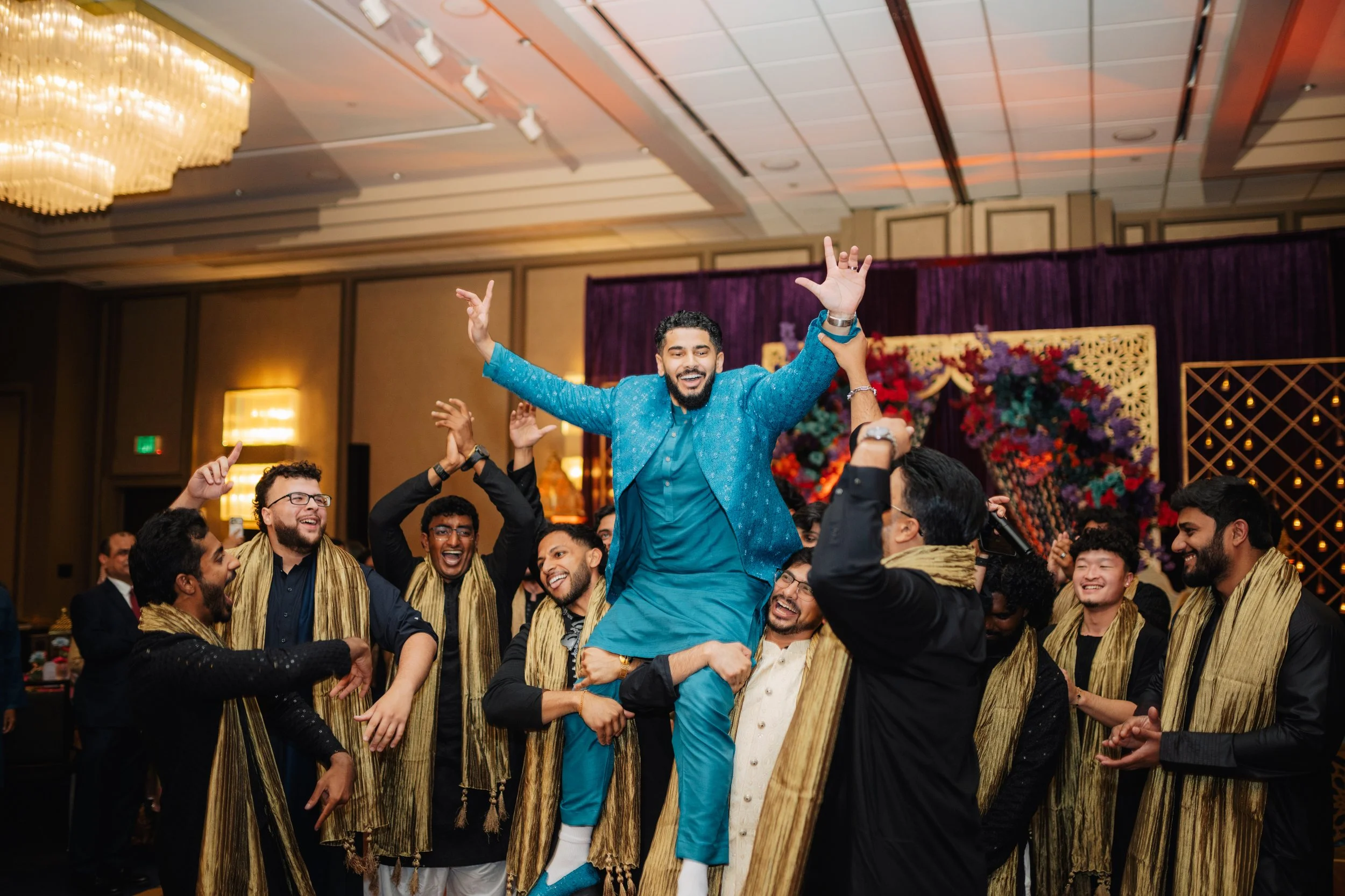 A group of men celebrating at a festive event, with one man being lifted in the air and everyone smiling and enjoying the moment.
