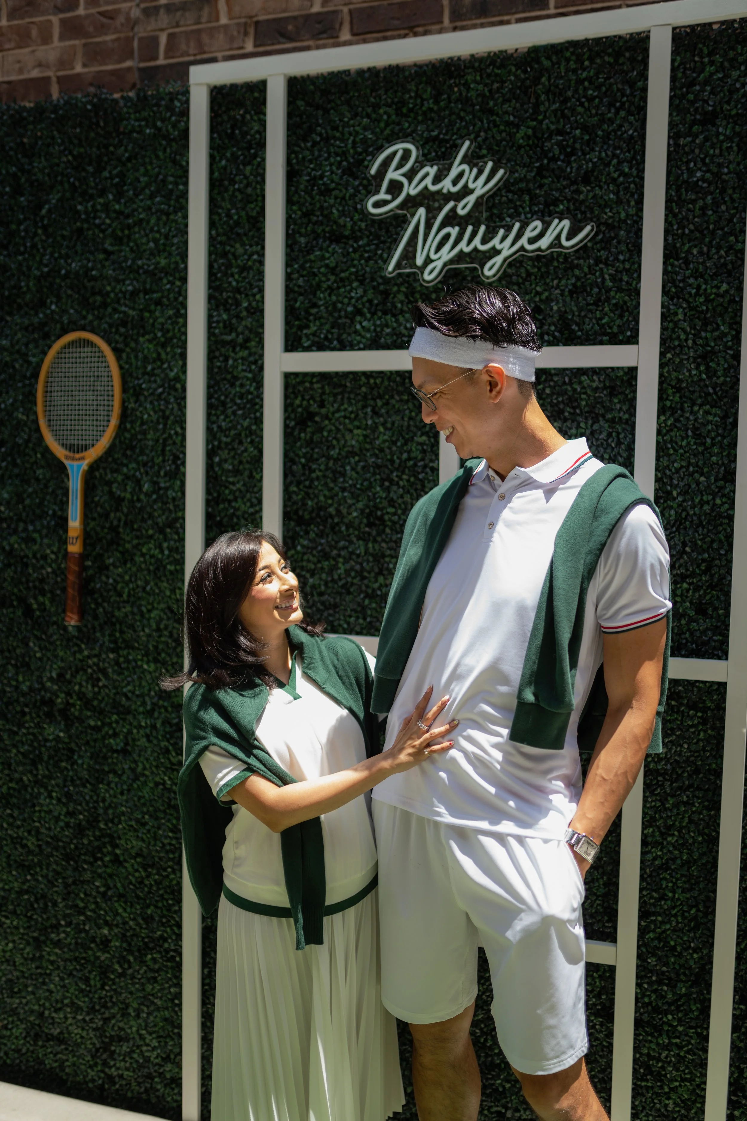 A woman and a man standing close together outdoors, smiling at each other. The woman is wearing a green and white athletic jacket with white pleated pants, and the man is wearing a white polo shirt with a green sweater draped over his shoulders and w