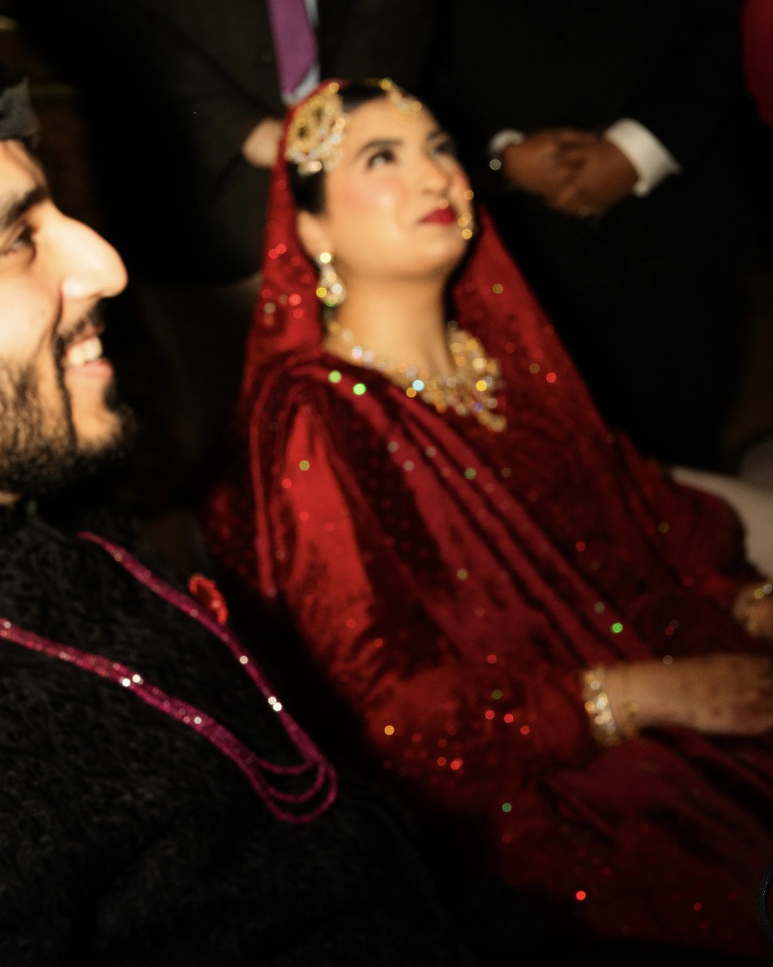 A woman in traditional Indian attire, wearing a red bridal dress with gold jewelry and a veil, seated beside a man wearing a dark outfit with pink accents.