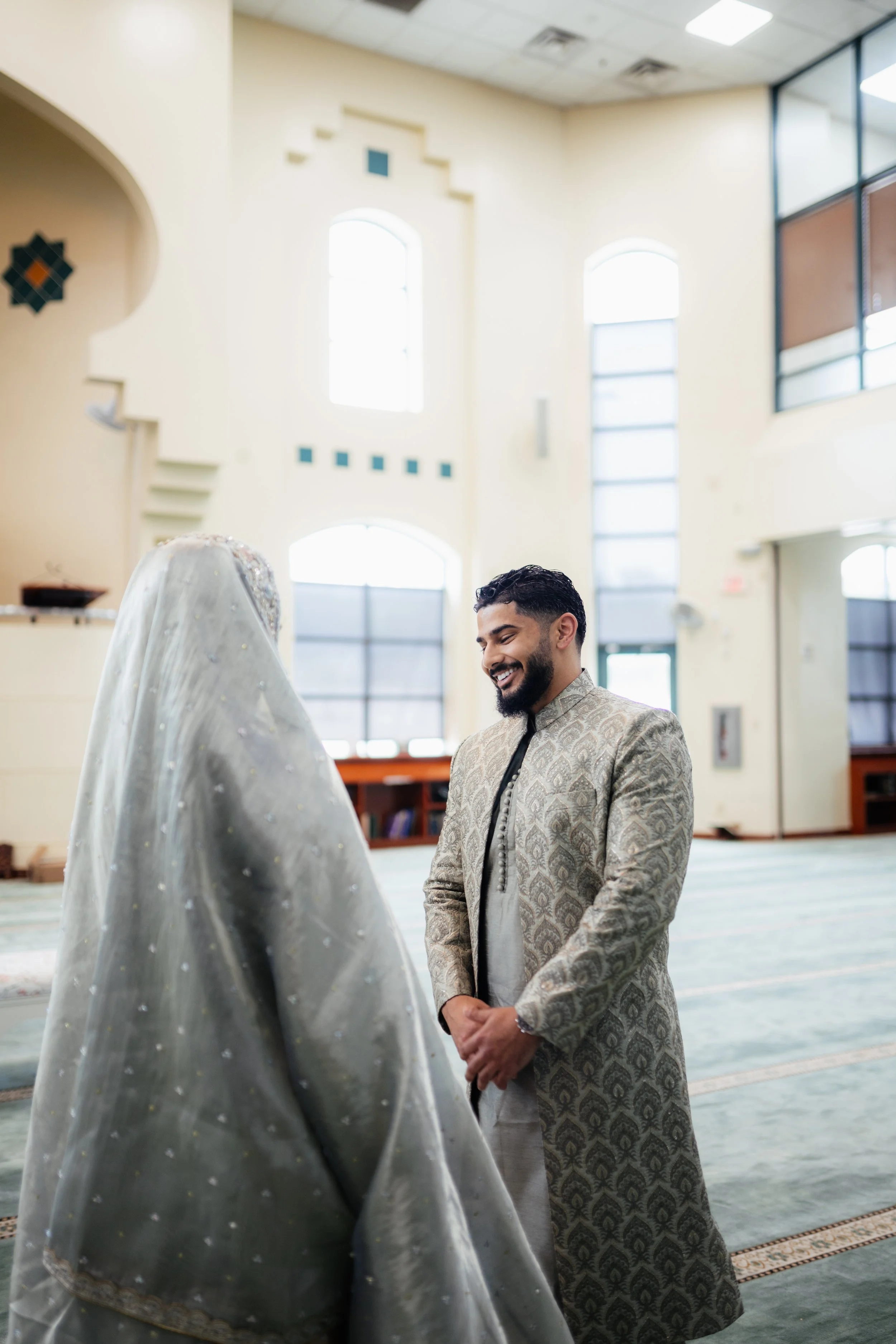 A man dressed in traditional South Asian attire smiling and standing in a spacious room with high ceilings and large windows, engaging in a moment of happiness or celebration.