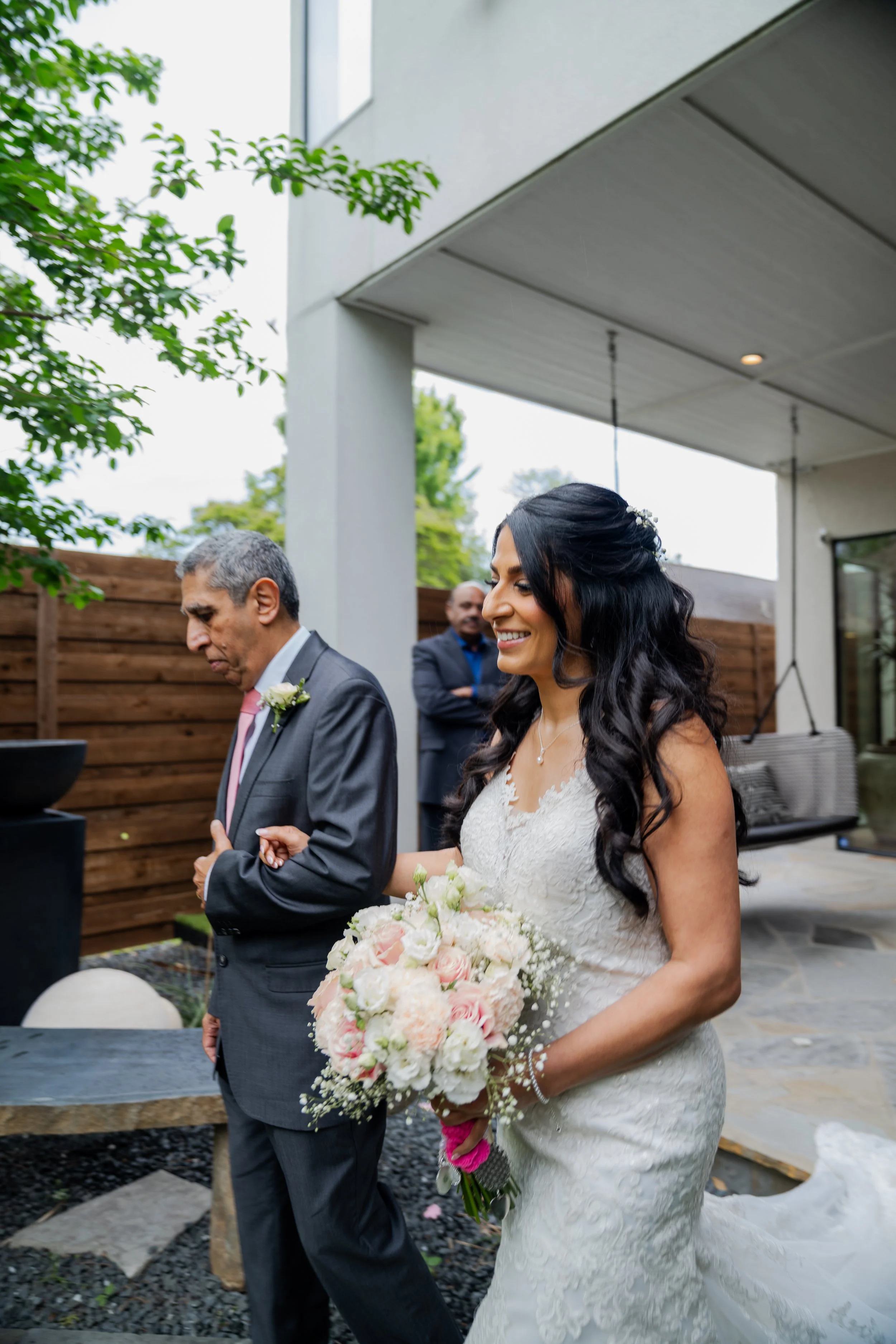 A bride smiling as she walks outdoors holding a large bouquet of pink and white flowers, accompanied by an older man in a suit, with a garden and a wooden fence in the background.
