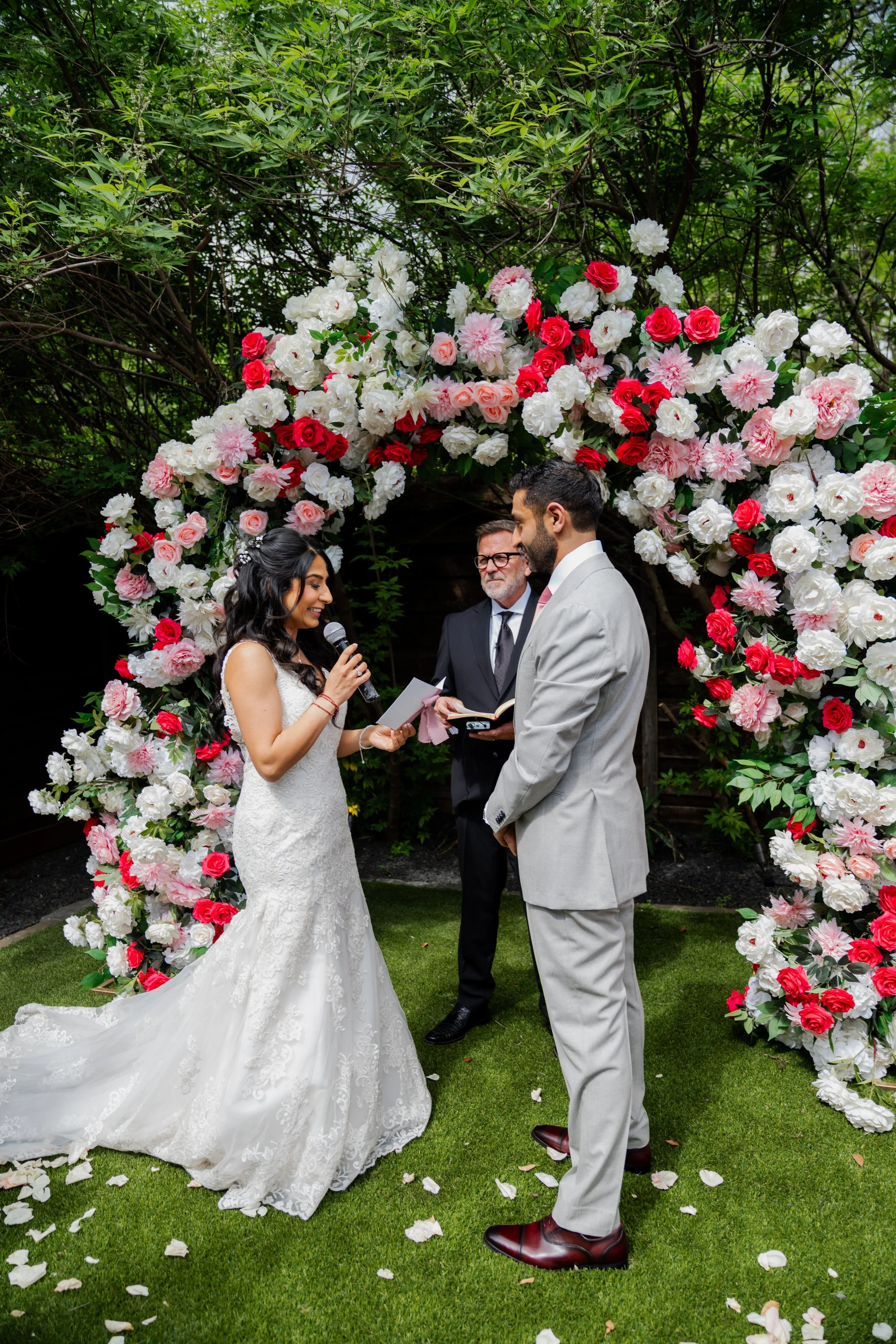 A bride and groom exchanging vows beneath a floral arch at an outdoor wedding ceremony. The bride wears a white lace gown, and the groom a light gray suit. An officiant stands between them holding a book. The scene is decorated with pink, white, and 