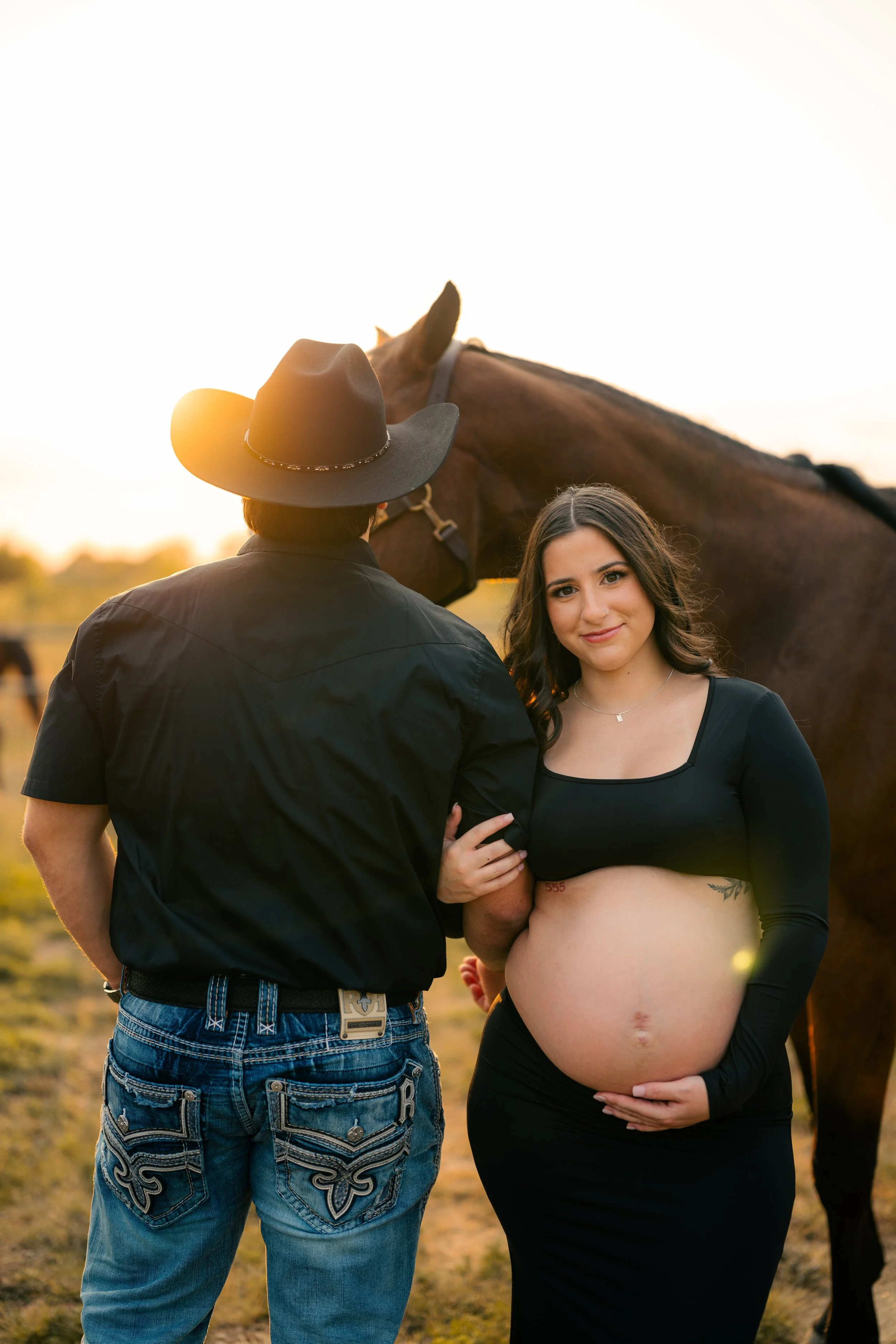 A pregnant woman holding her belly standing next to a man in a cowboy hat and black shirt, with a horse nearby at sunset.