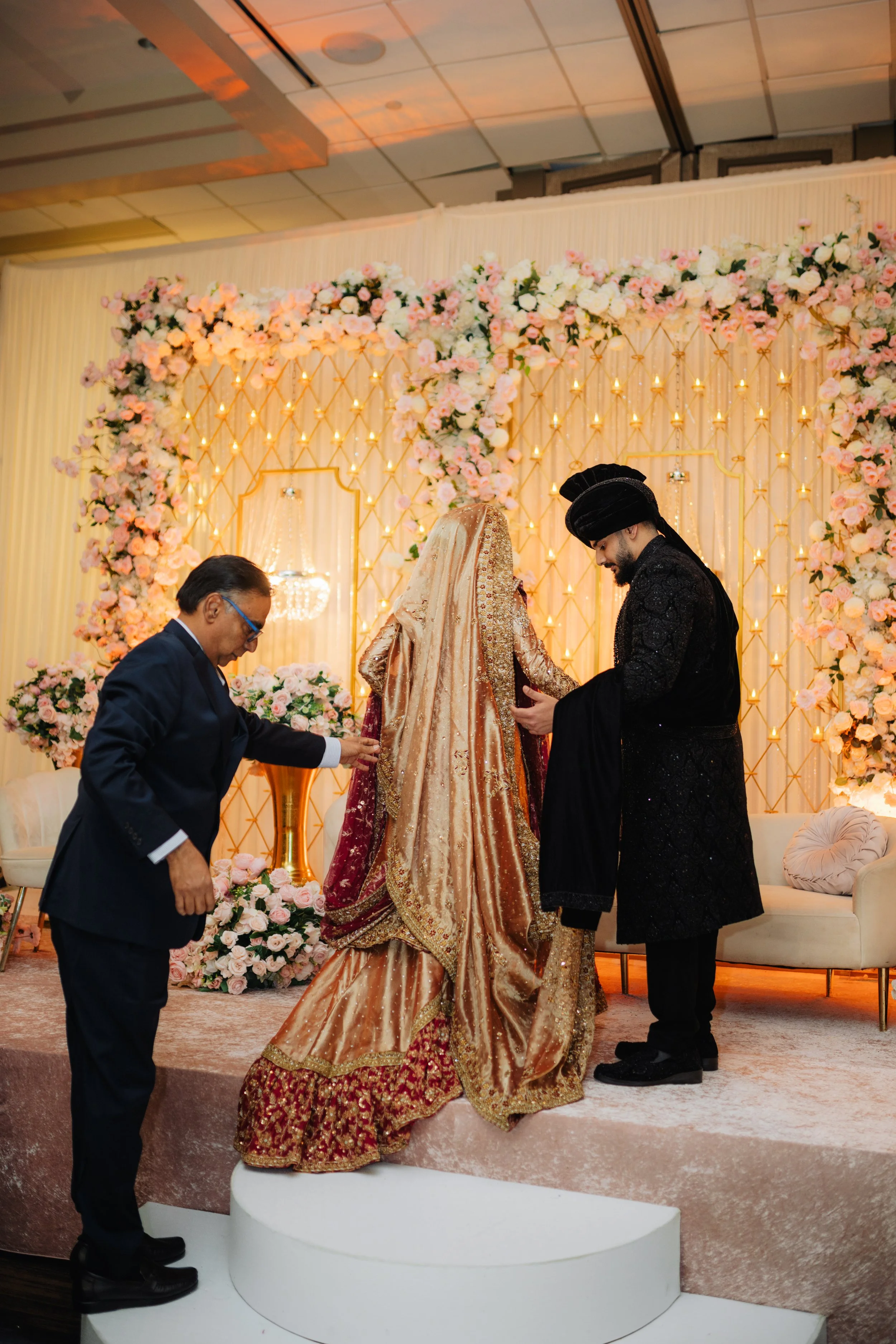 A traditional wedding ceremony with a bride in an ornate gold and red gown, standing on a white platform, exchanging vows with a groom dressed in a black embroidered sherwani and turban. An officiant and another man in formal attire assist, with a ba