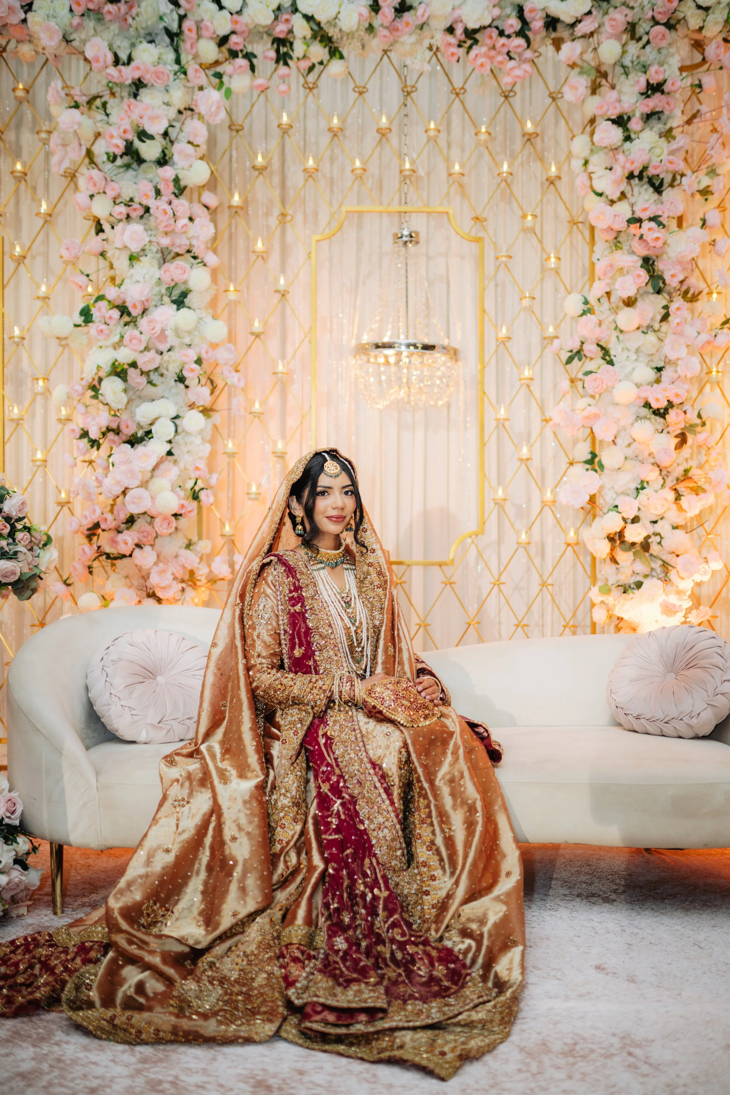 A woman dressed in traditional South Asian bridal attire sitting on a white sofa in front of a floral backdrop with pink and white roses and gold accents.