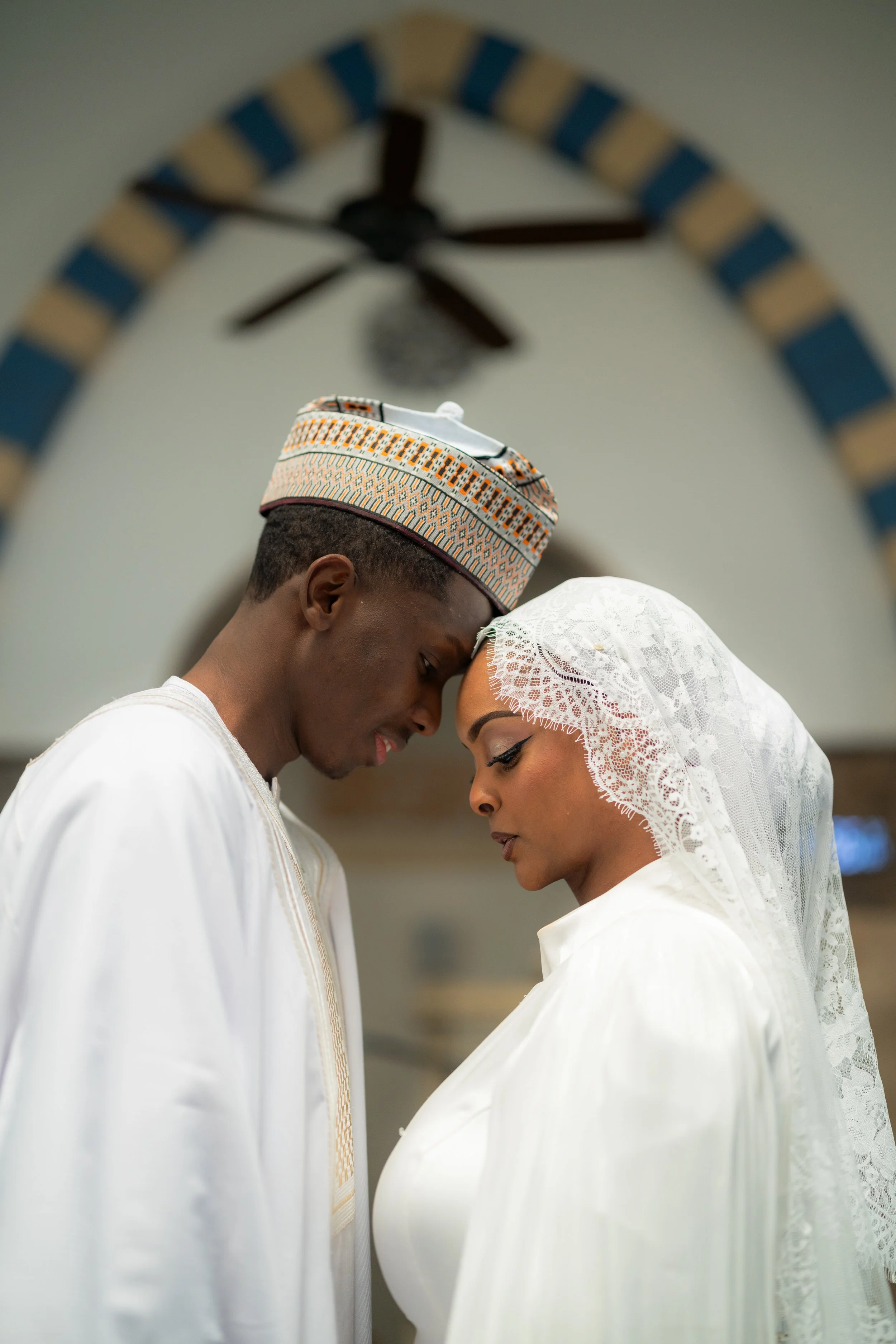 A Muslim couple in traditional white attire and head coverings sharing a tender moment indoors, with their foreheads touching.
