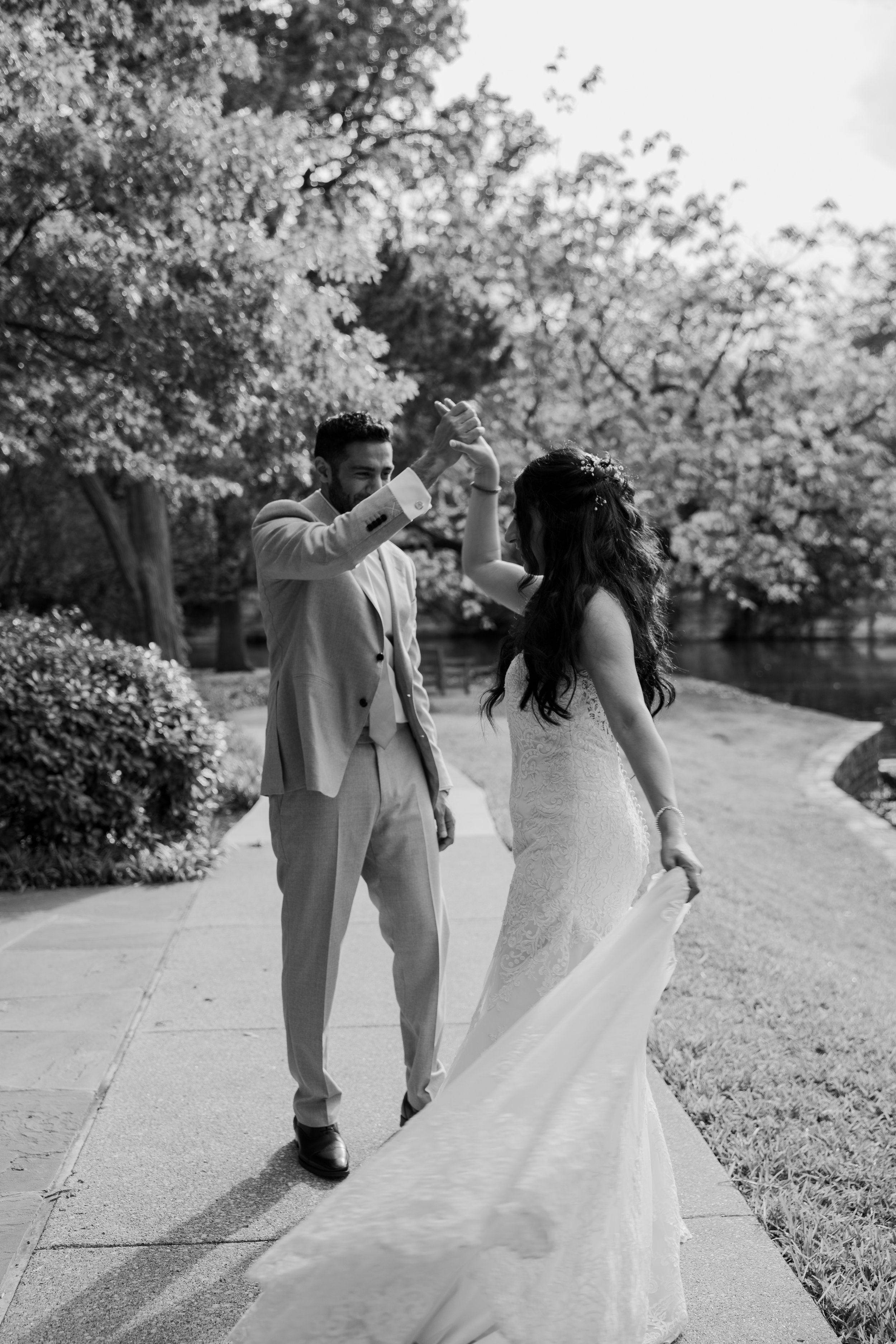 A black and white photo of a bride and groom dancing outdoors, with the groom in a light-colored suit and the bride in a lace wedding gown. The groom holds the bride's hand, and the bride holds a hat in her other hand. They are on a sidewalk, surroun