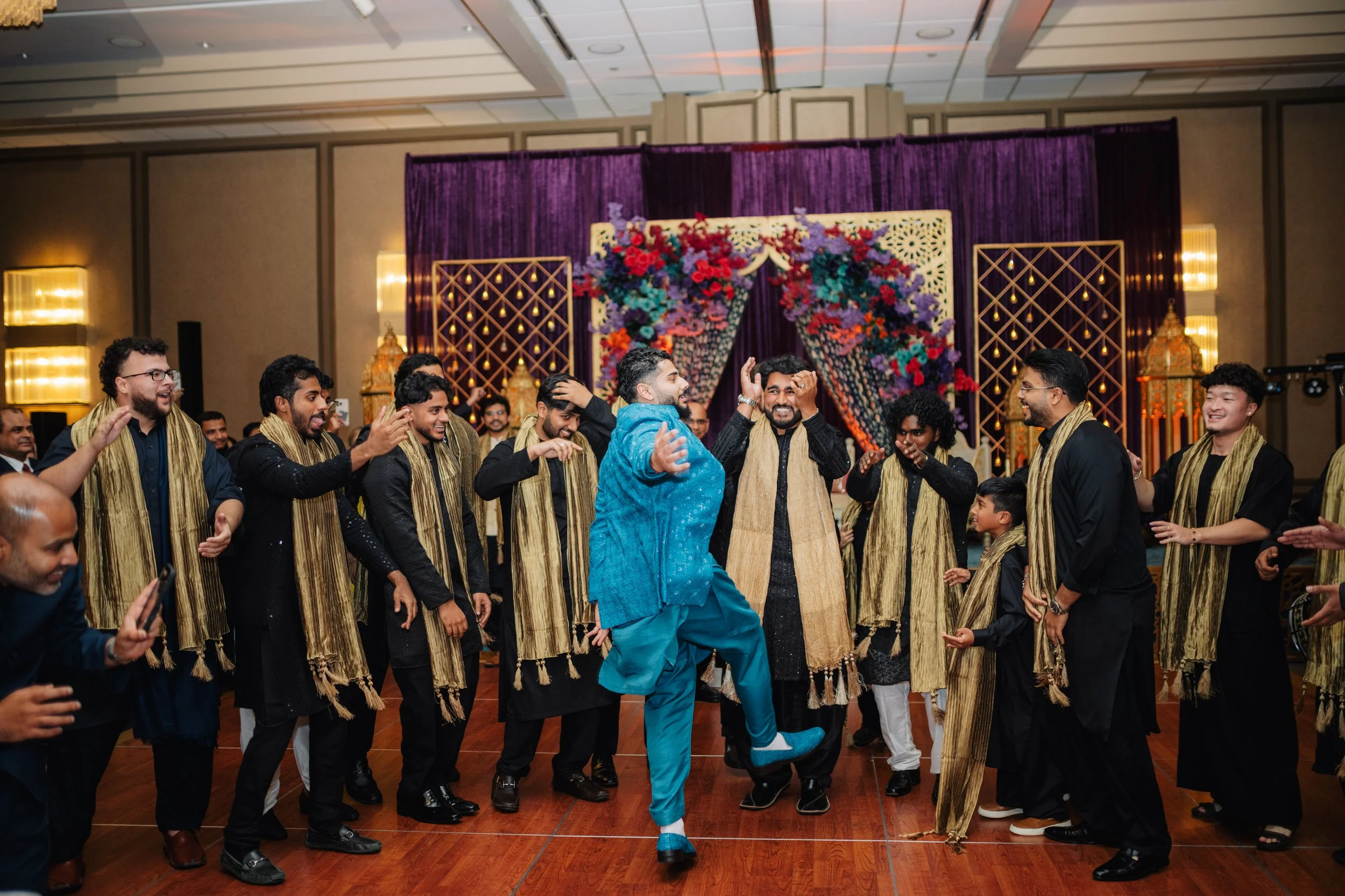 Group of people dressed in traditional Indian attire dancing and celebrating at an indoor event with a floral backdrop.