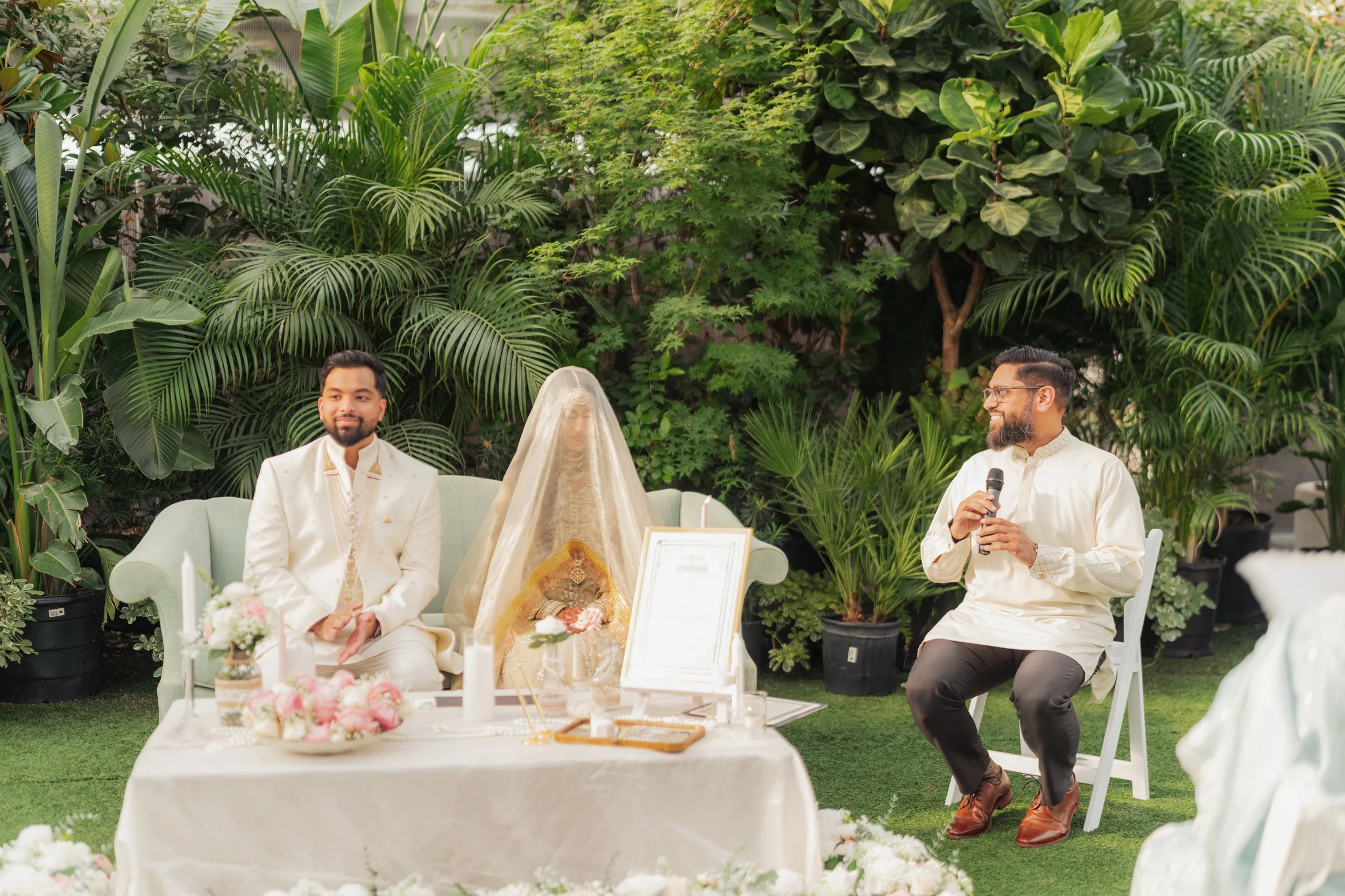 A wedding ceremony with a bride and groom seated outdoors, surrounded by lush green plants, with a man standing and speaking into a microphone.