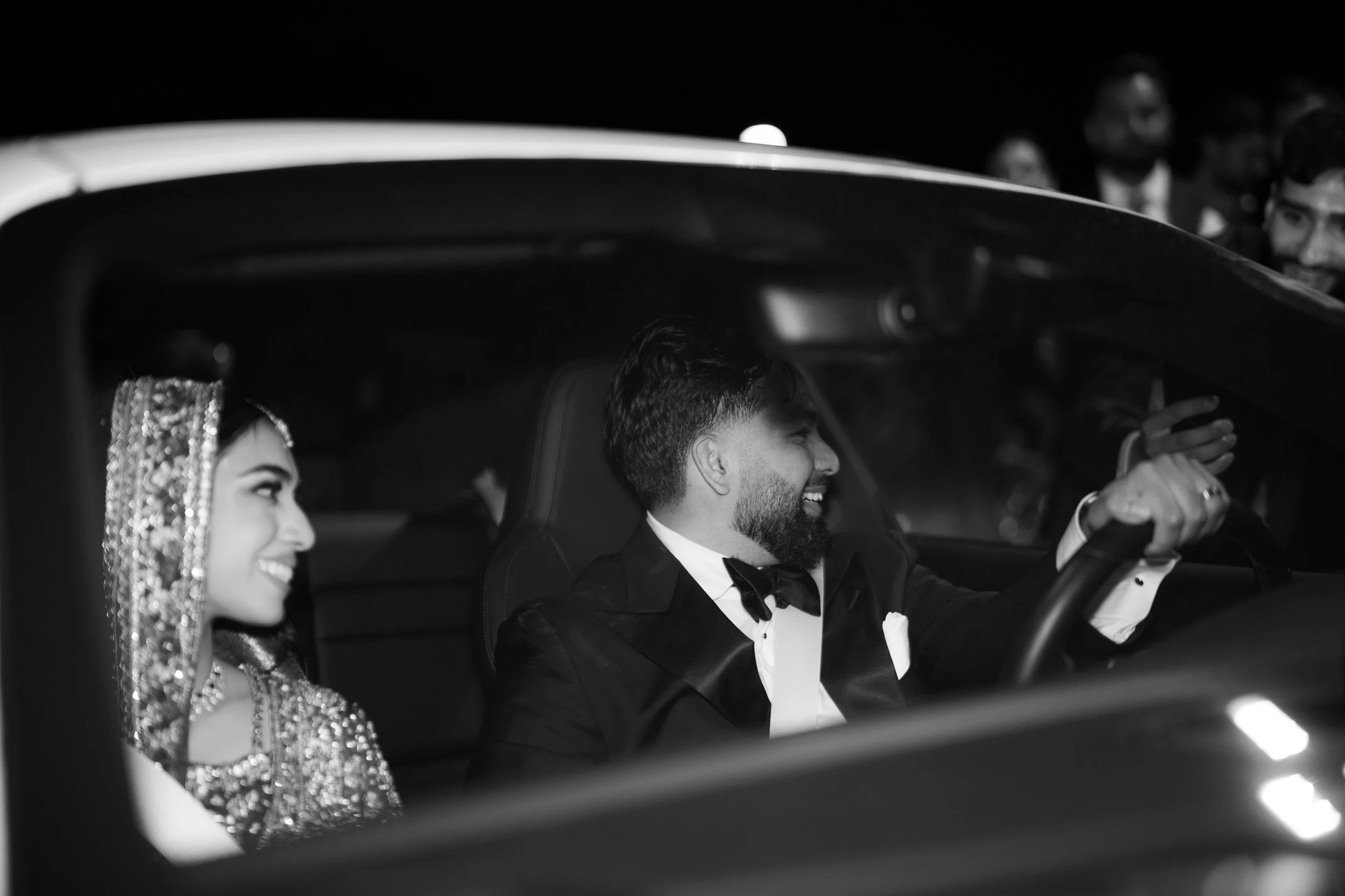 A black and white photo of a bride and groom in a car at night, smiling. The bride is wearing a bejeweled headpiece, and the groom is dressed in a tuxedo. There are people in the background witnessing the moment.