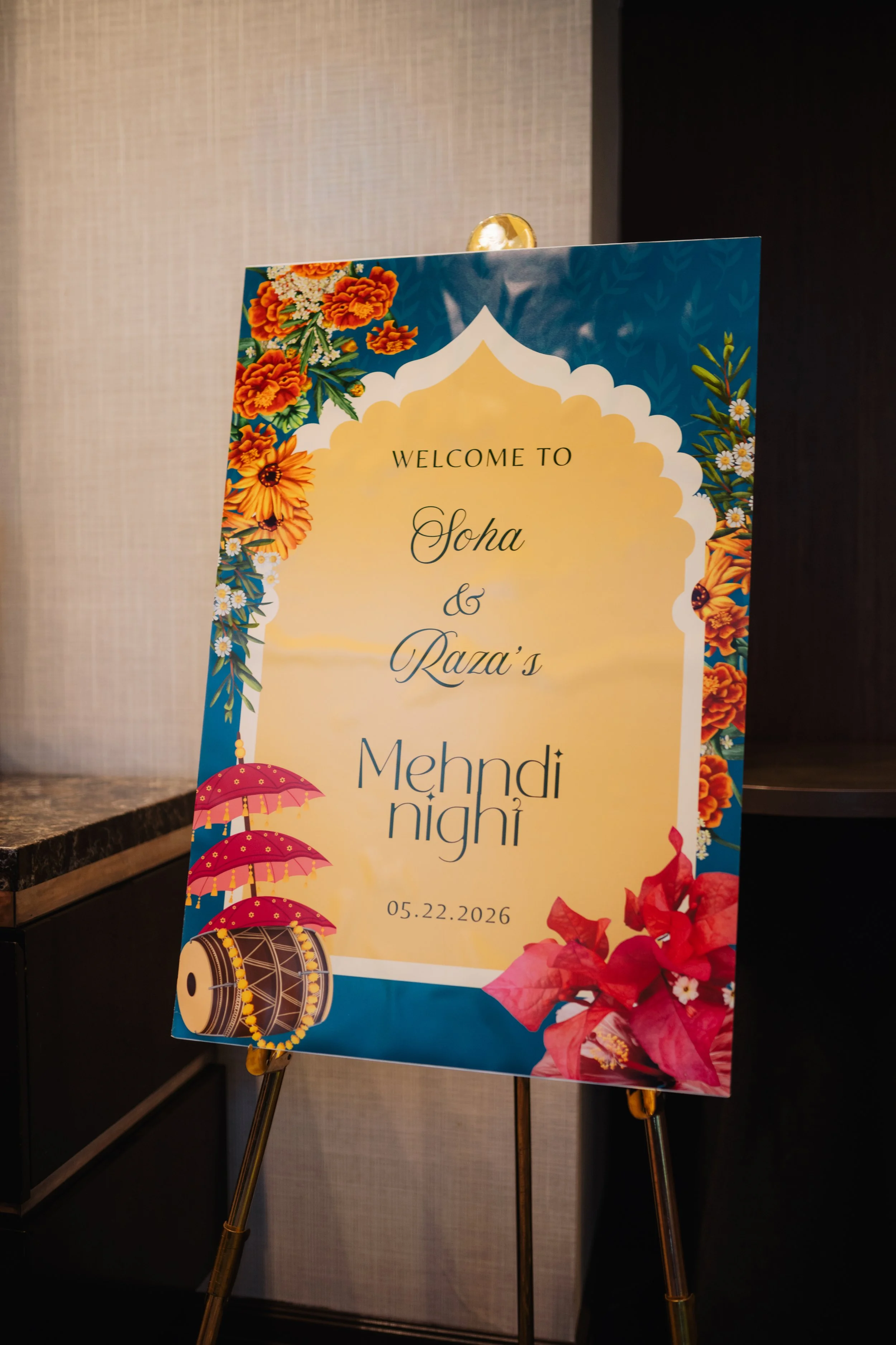 Colorful welcome sign with flowers, umbrellas, a drum, and a date, indicating a celebration or event.