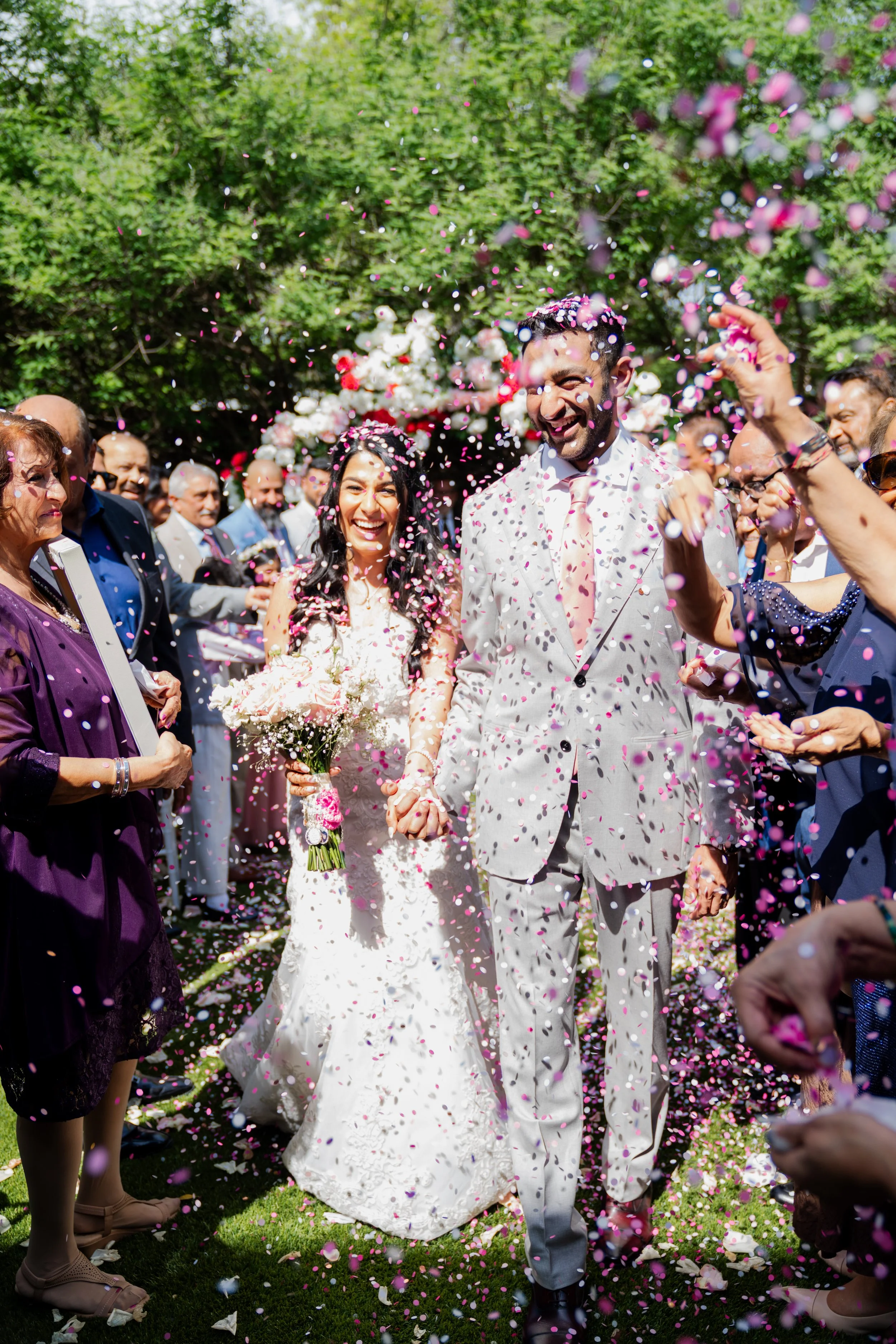 A wedding celebration outdoors with a bride and groom holding hands and smiling, surrounded by guests throwing pink and white confetti, with greenery and flowers in the background.