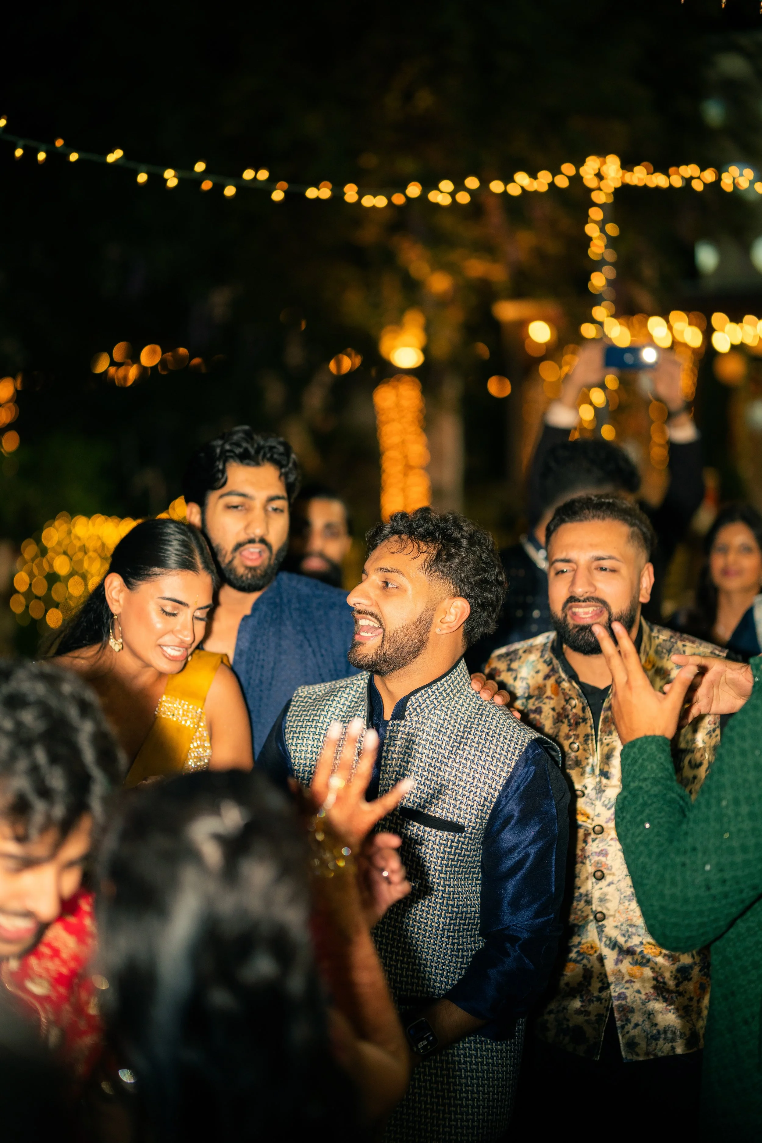 People wearing traditional Indian attire celebrating at a nighttime outdoor party with string lights.