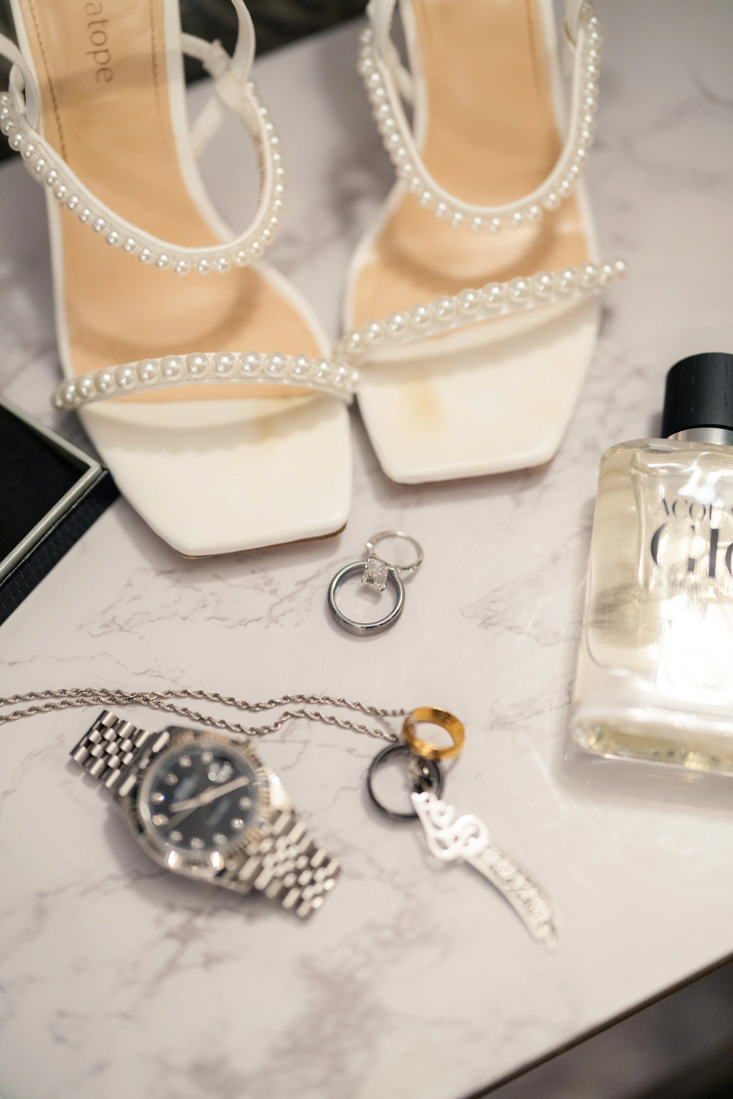 White high-heeled shoes with pearl embellishments near toes, a bottle of perfume, a silver watch, and wedding rings on a white marble surface.