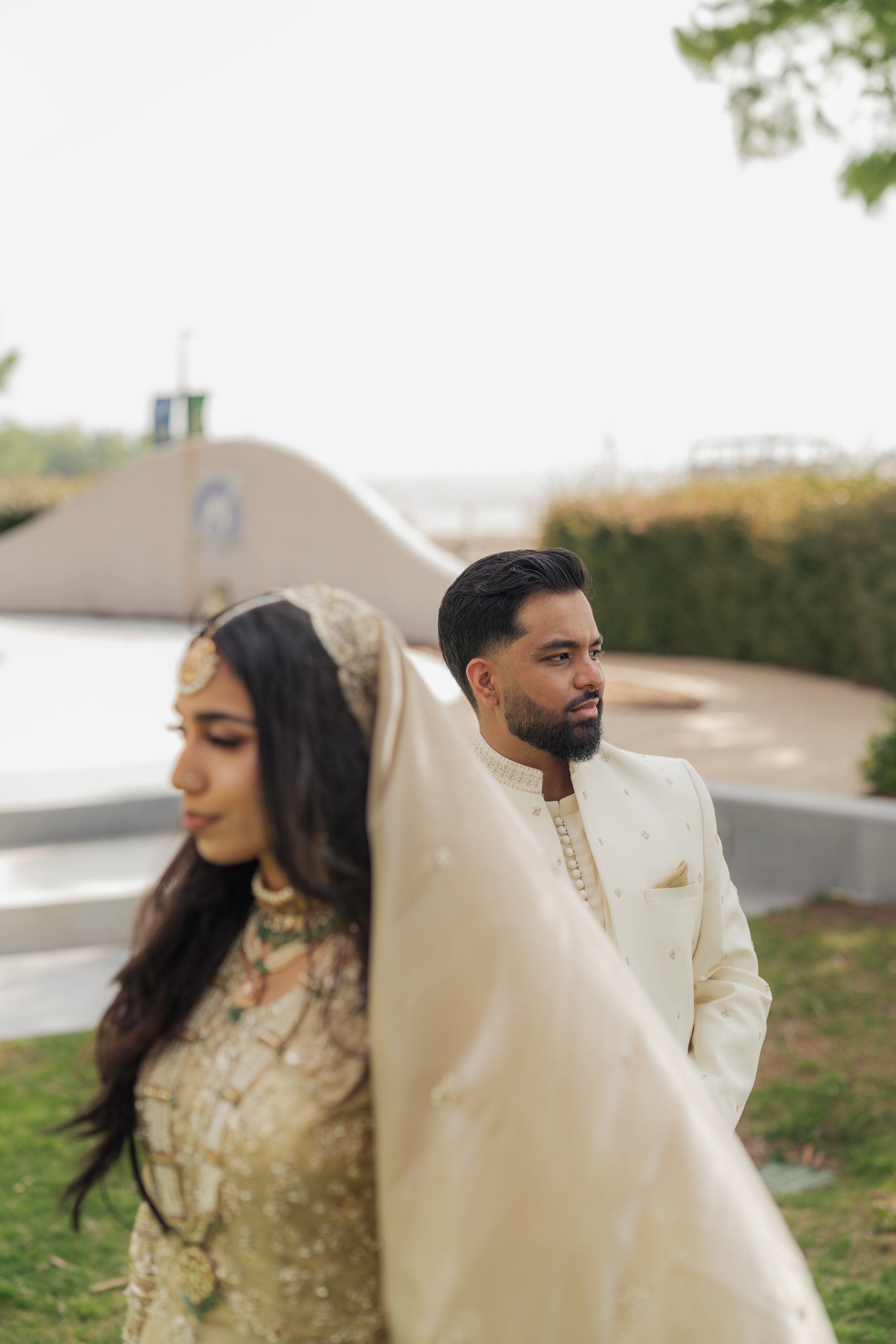 A bride and groom standing outdoors on their wedding day, dressed in traditional Indian attire. The bride has long dark hair and is wearing a gold and green embroidered saree with jewelry, and the groom has short black hair and a beard, wearing a cre