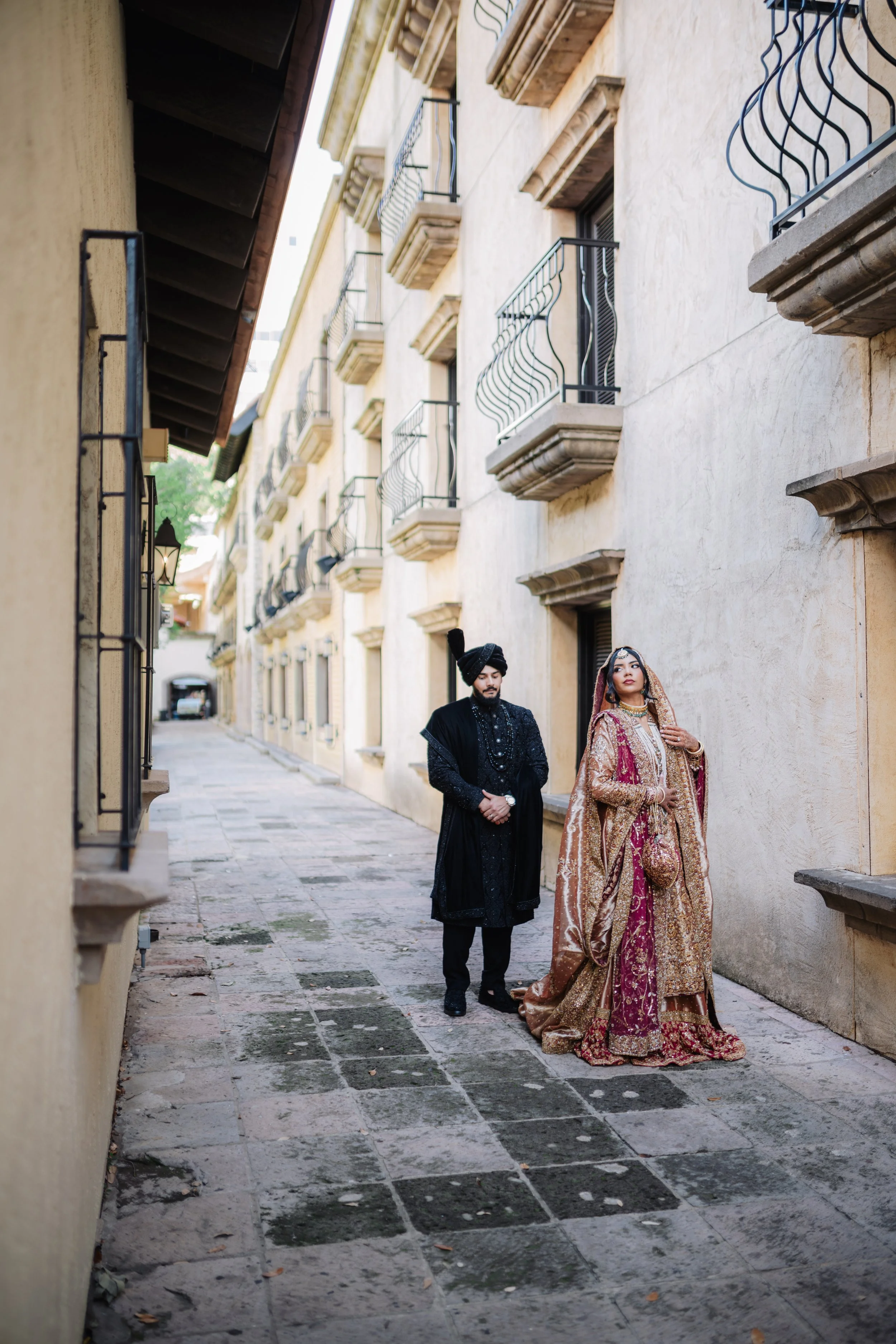 A man and woman dressed in traditional South Asian clothing walking down a narrow stone alleyway with beige walls and balconies on one side.