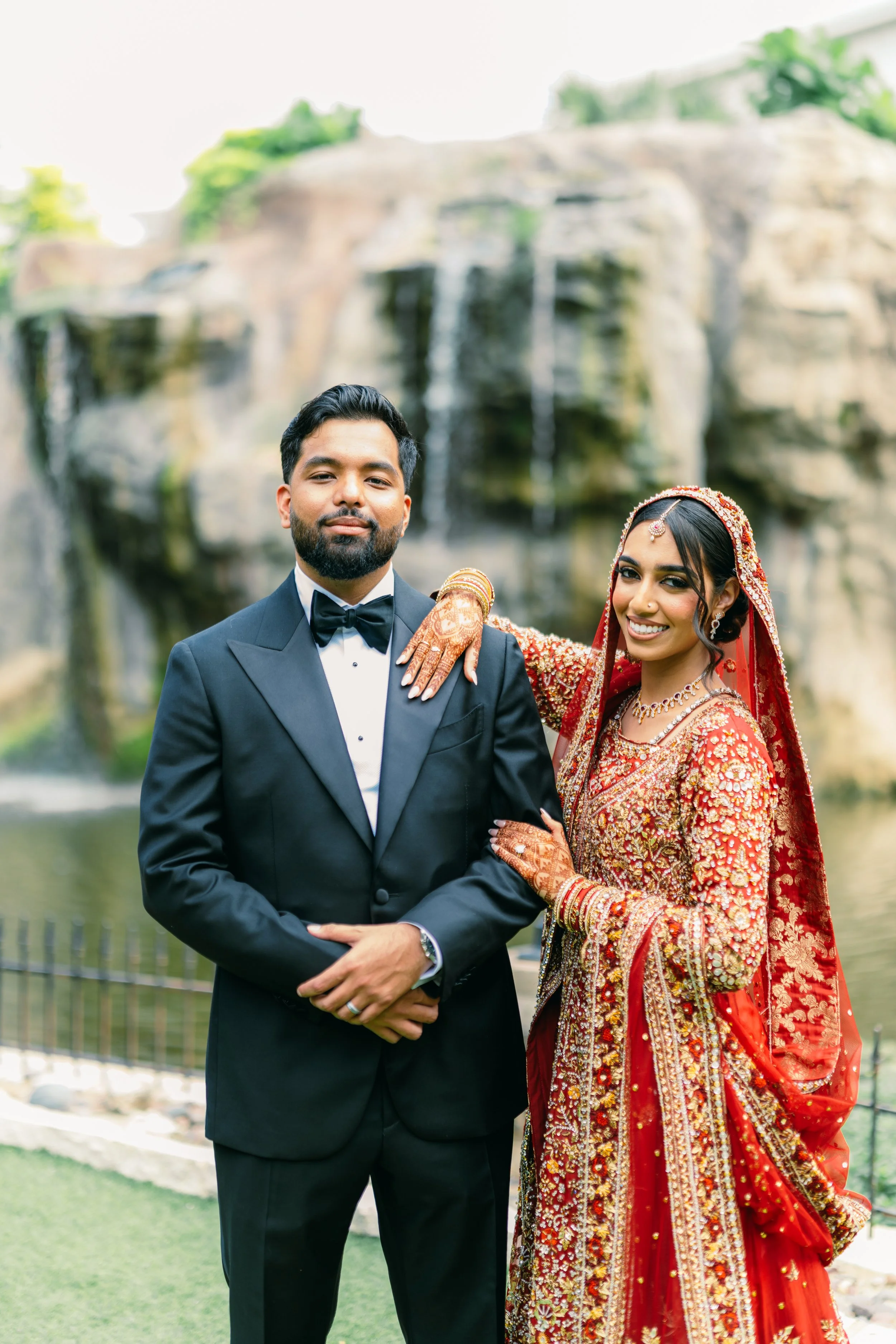 A couple dressed in traditional Indian wedding attire outdoors, with a waterfall and greenery in the background.