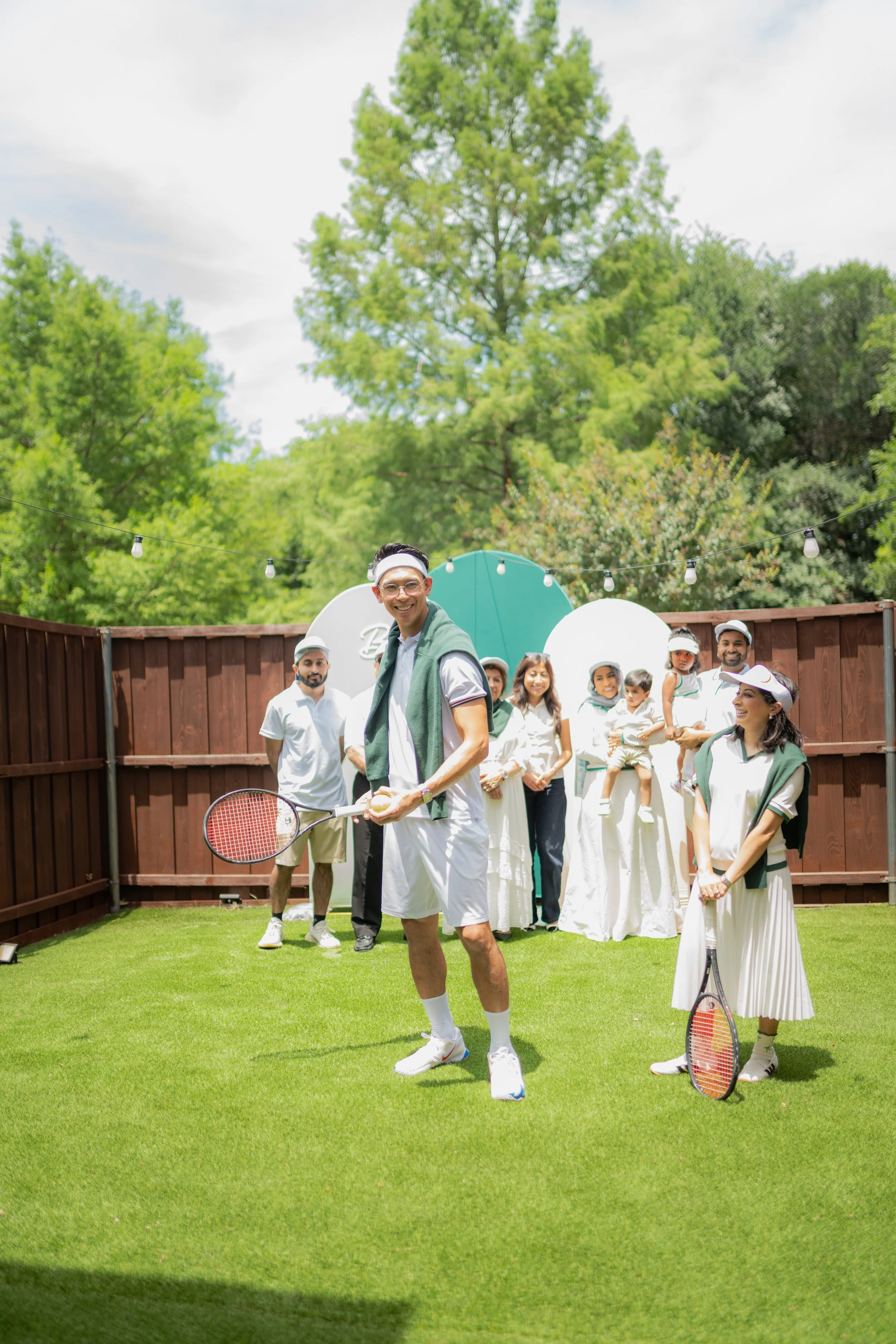A group of people at a backyard gathering, some holding tennis rackets, on green grass with trees and string lights in the background.