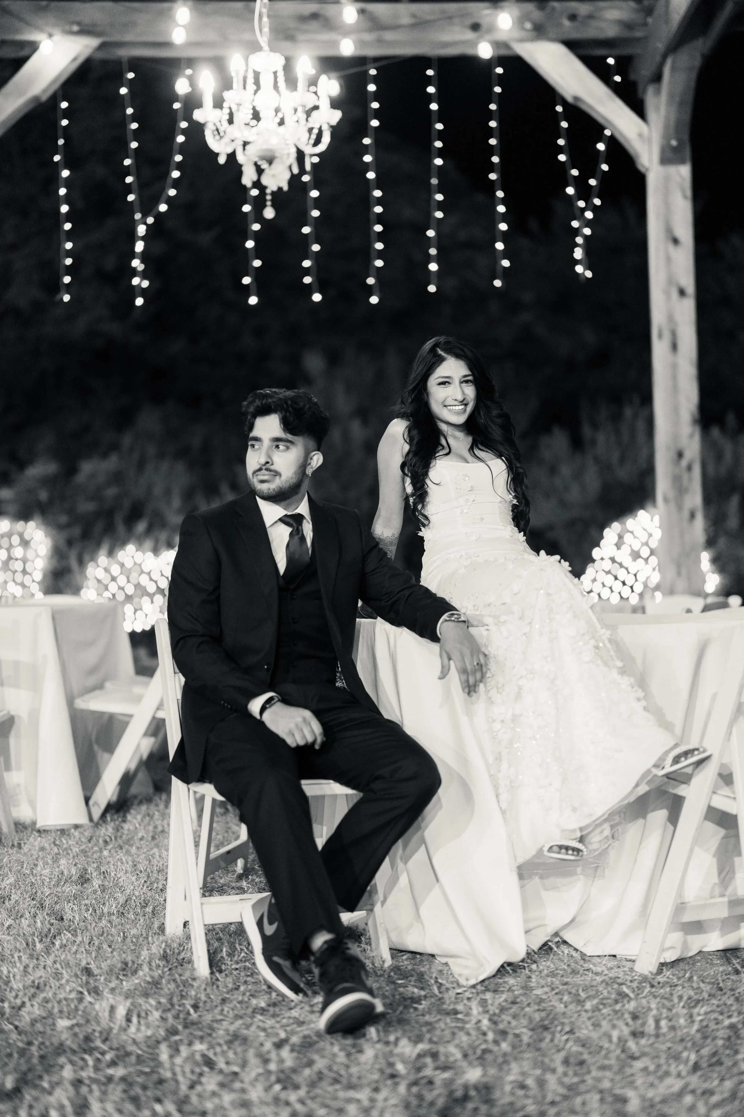 A black and white photo of a couple at a wedding reception, with the bride in a wedding dress and the groom in a suit, sitting on chairs under a wooden pavilion decorated with hanging string lights and a chandelier.