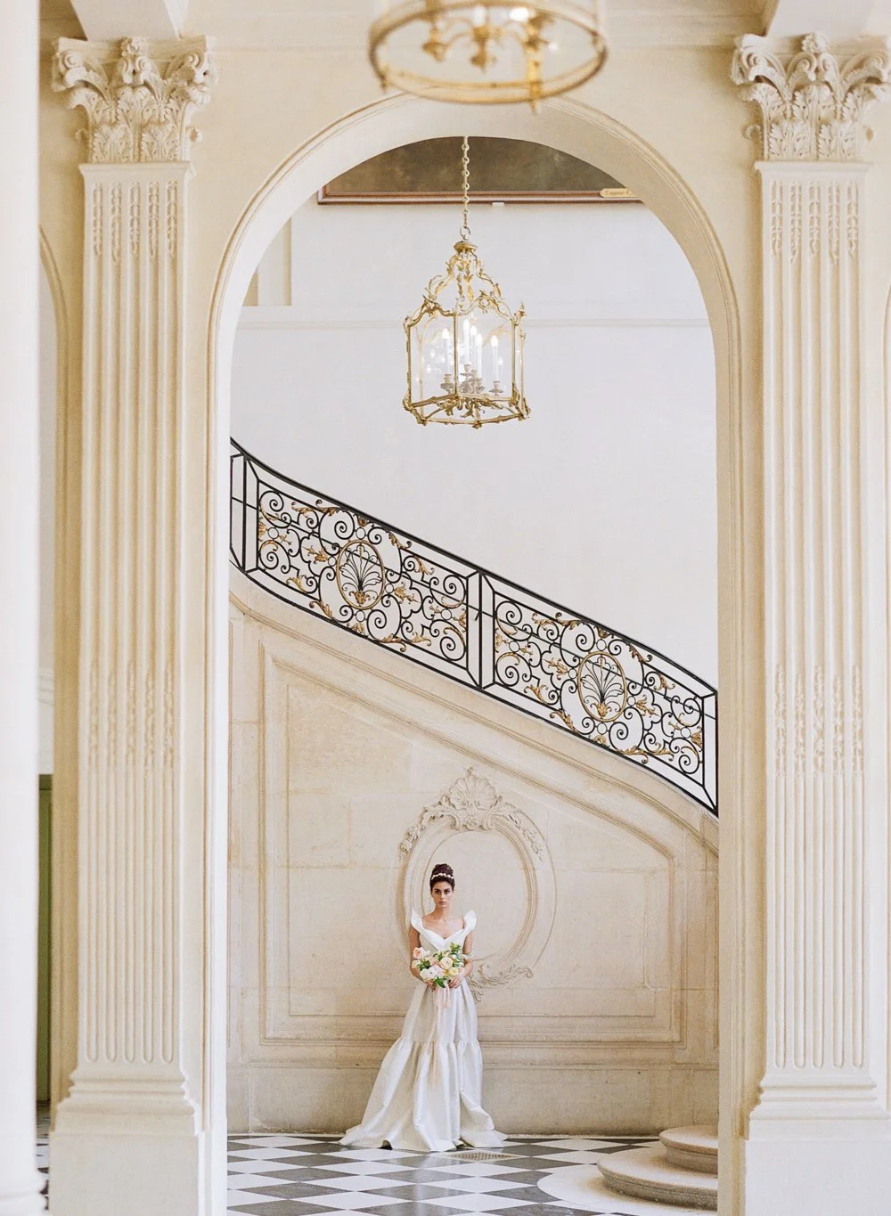 Elegant interior with a woman in a white gown holding flowers, standing by a staircase with ornate iron railing, flanked by classical columns and a chandelier.