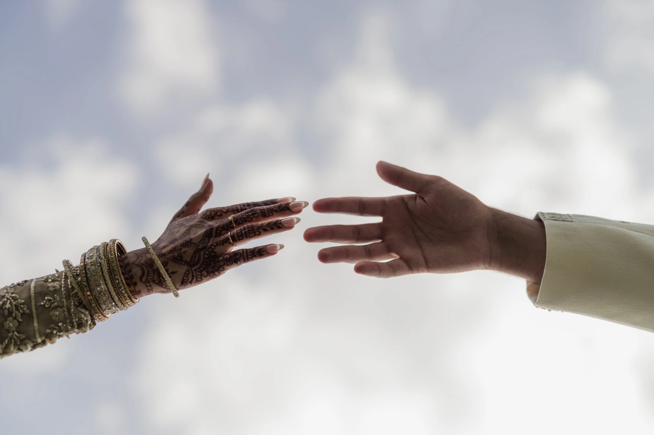 Close-up of two hands reaching towards each other against a cloudy sky, one decorated with jewelry and traditional henna designs, and the other in a white sleeve.