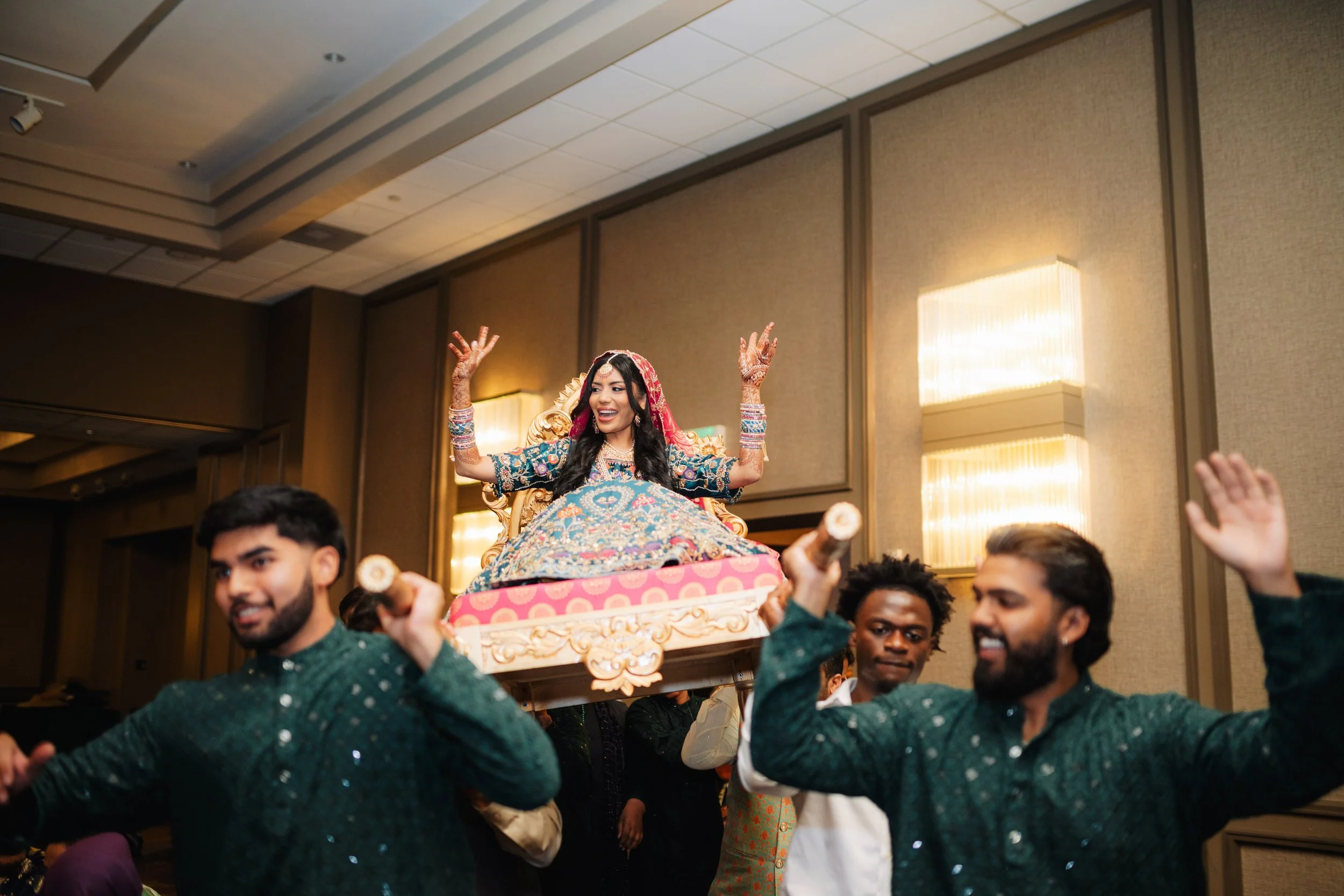 A young woman dressed in traditional Indian attire sits on a decorated throne being carried on a platform by a group of men during a festive celebration indoors.