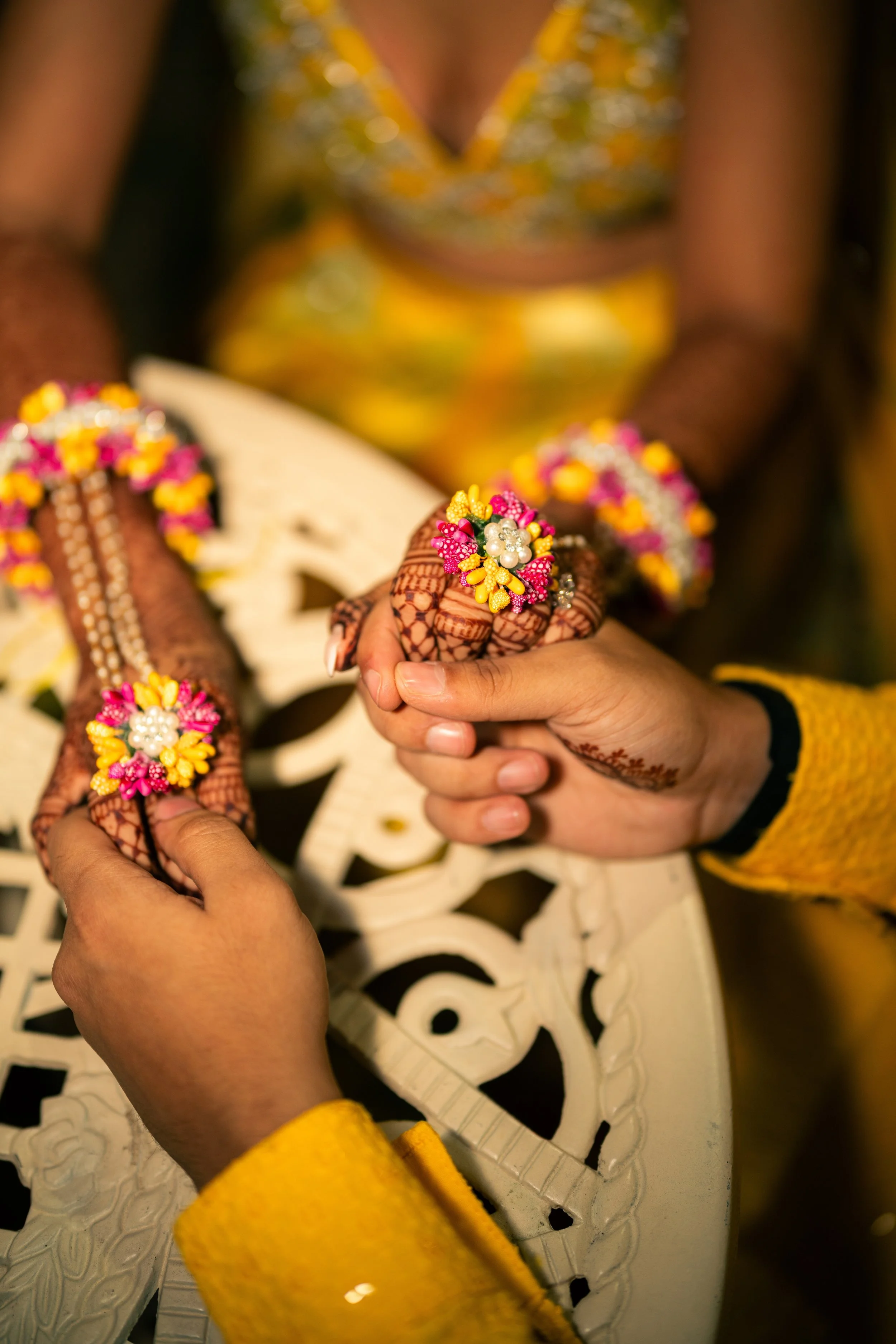 Close-up of two hands holding each other, decorated with vibrant floral jewelry, with a blurred woman in a yellow sari in the background.