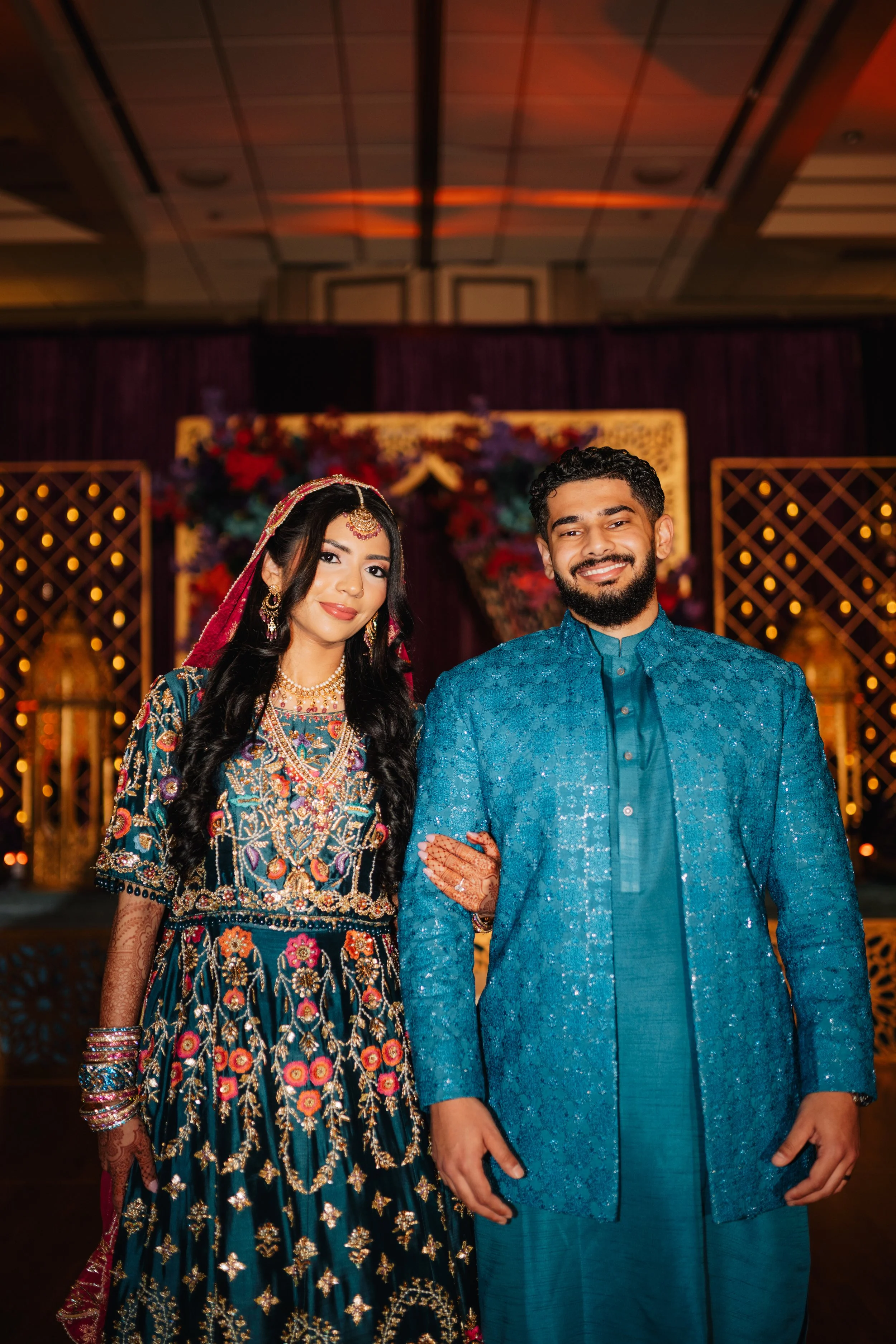 A South Asian couple dressed in traditional Indian wedding attire, standing in front of a decorated backdrop with flowers and lights, smiling at the camera.