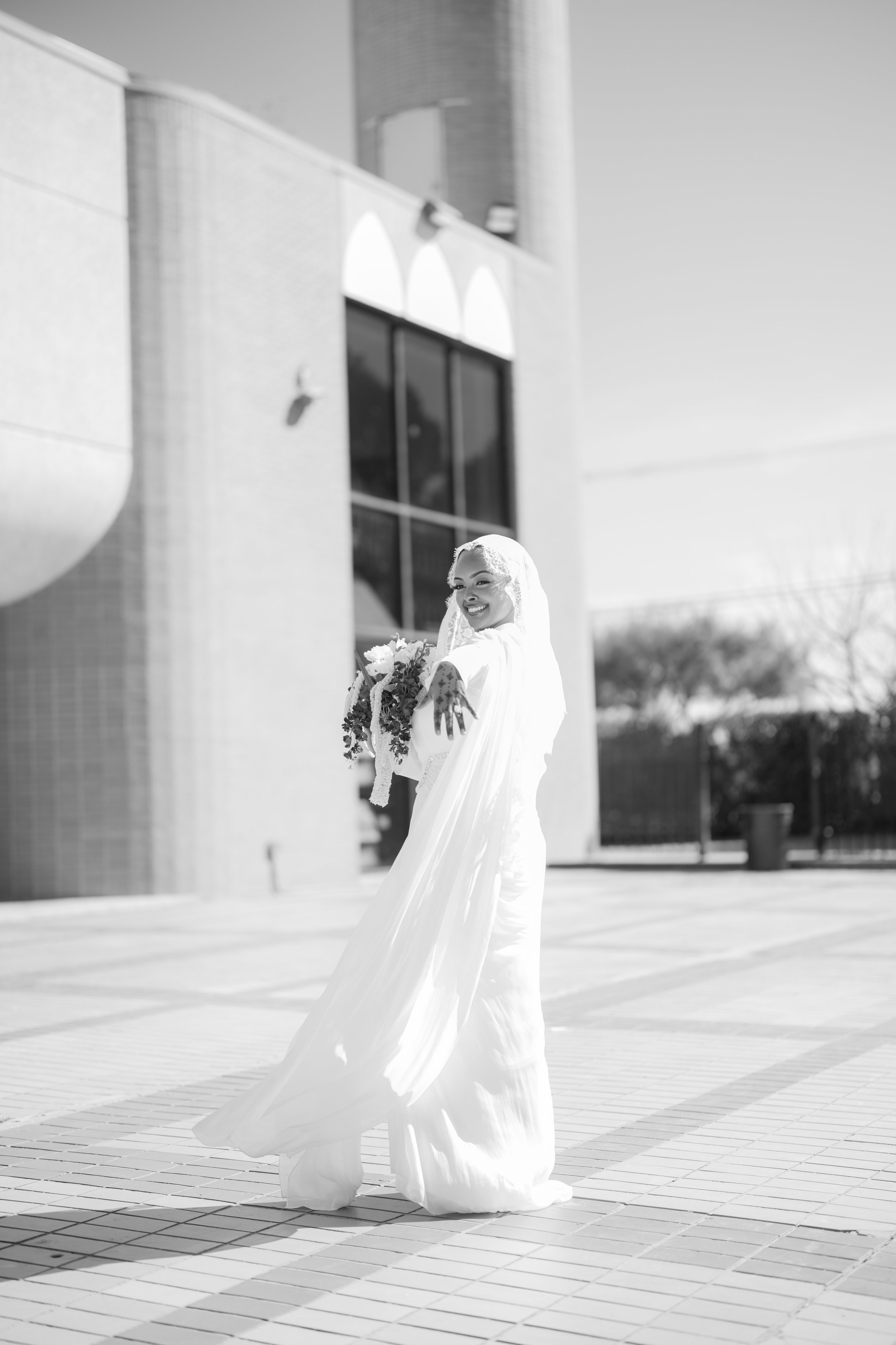 A woman in a wedding dress holding a bouquet, smiling outside on a sunny day.