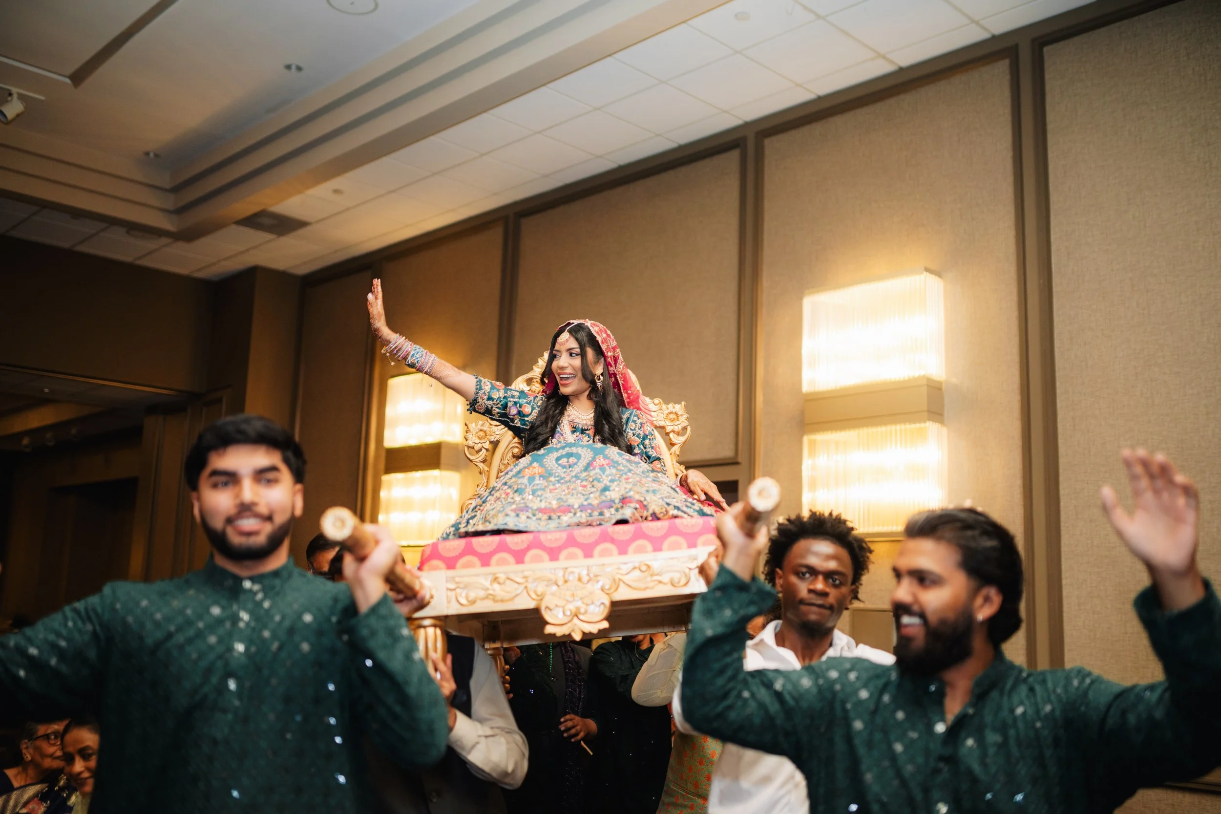 A woman in traditional Indian attire is being carried on a decorative palanquin during a celebration. She is smiling and waving, surrounded by joyful people.