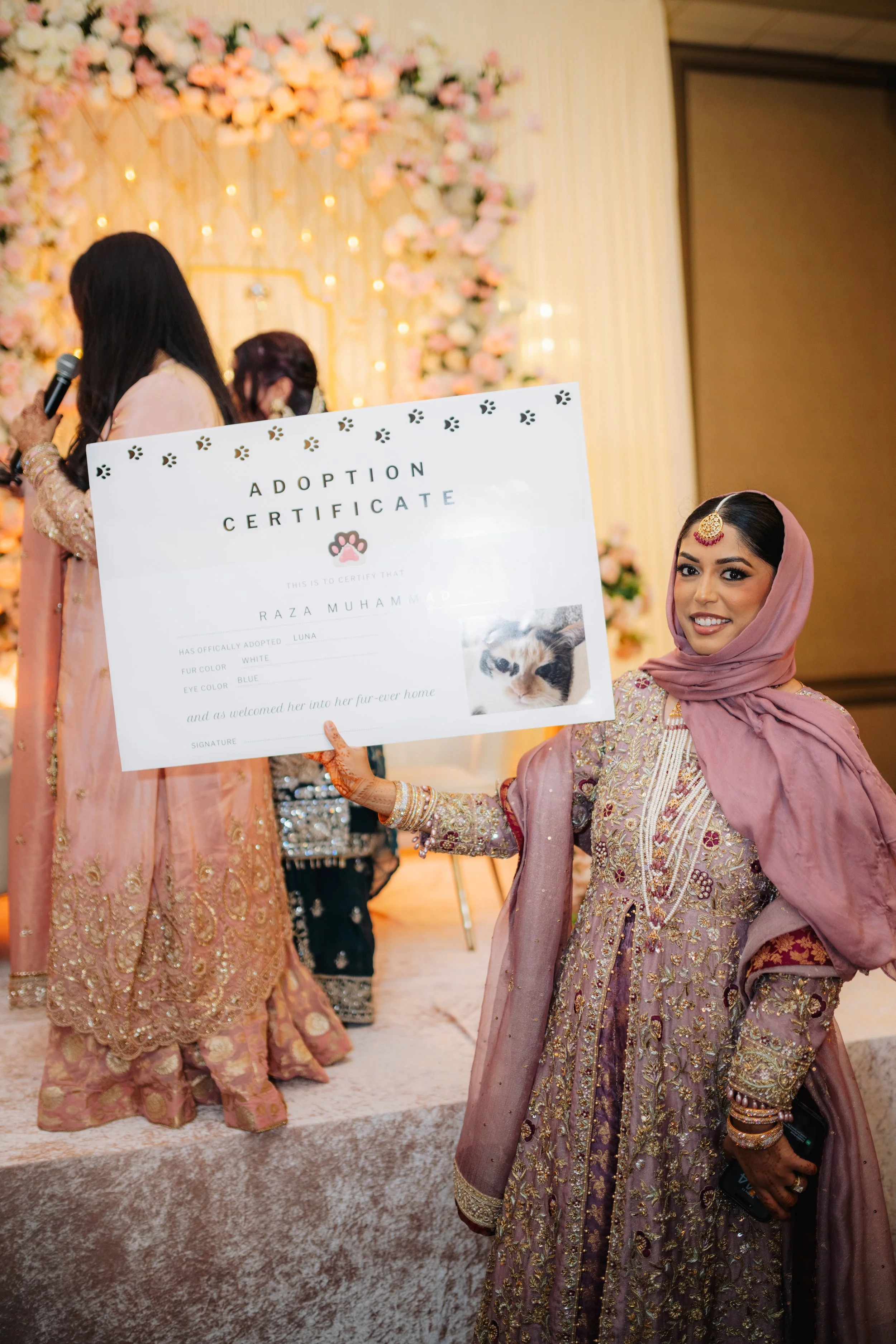 Woman in traditional attire holding an adoption certificate, standing in a decorated event hall with floral backdrop.
