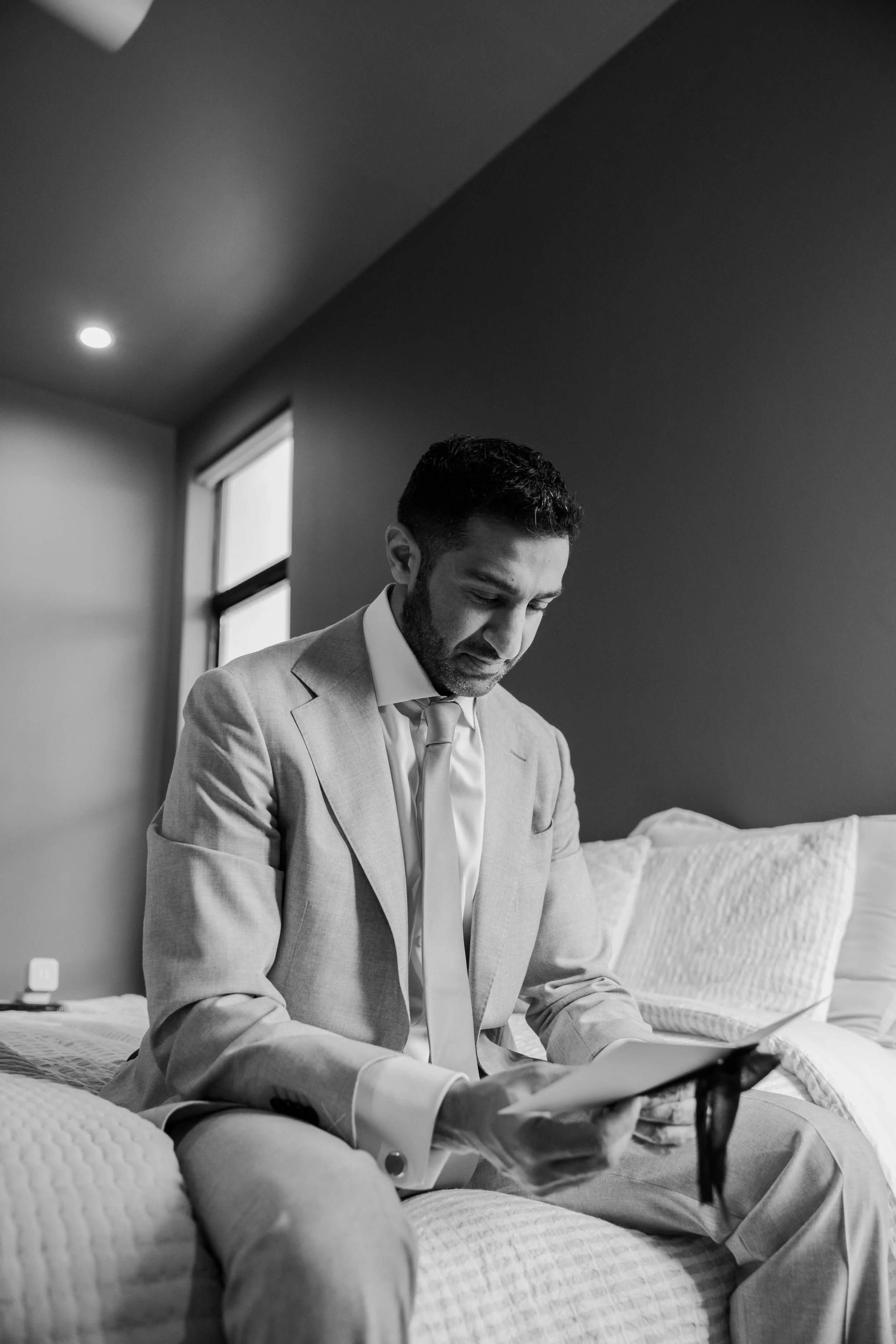 A man in a suit sitting on a bed, looking down at a tablet or book, in a bedroom with a window and minimal decor, black and white photograph.
