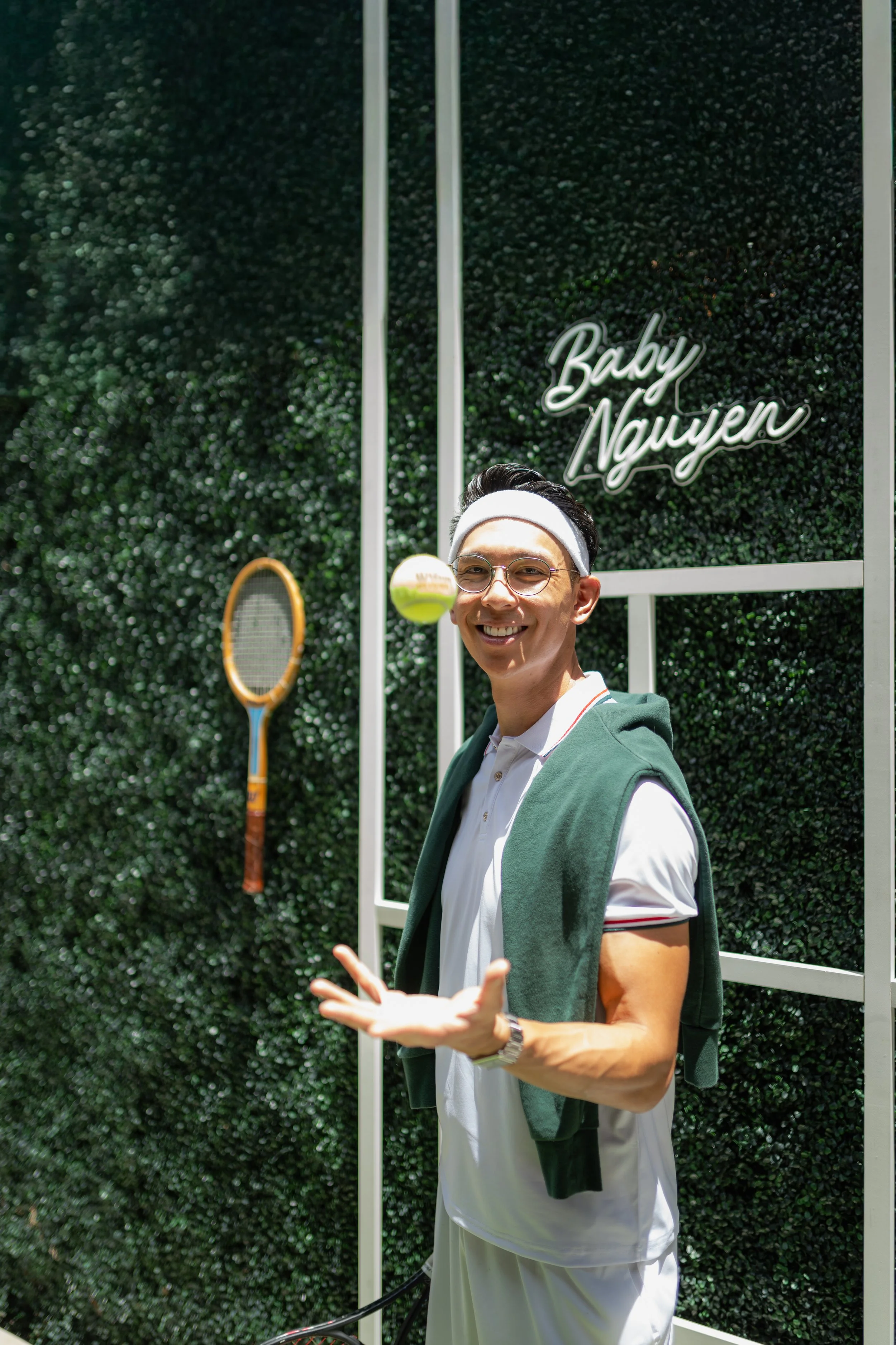 A young man smiling, wearing glasses, a white headband, a white polo shirt, and a green hoodie draped over his shoulders. He stands in front of a green hedge with a neon sign that reads 'Baby Nayun.' There is a tennis racket visible on the left side 
