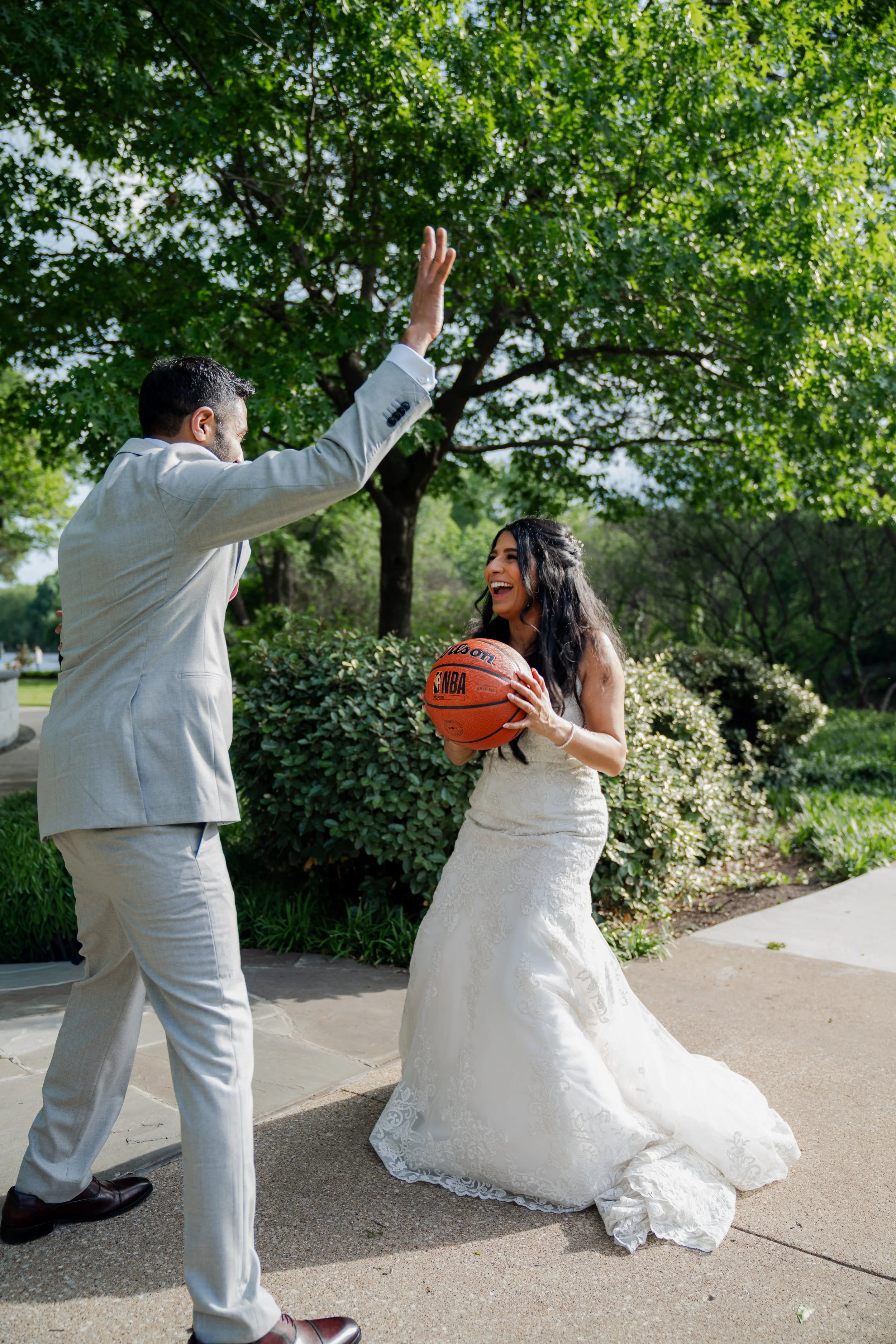 A man in a light gray suit and a woman in a wedding dress share a joyful moment outdoors, with the woman holding a basketball, under a green leafy tree.