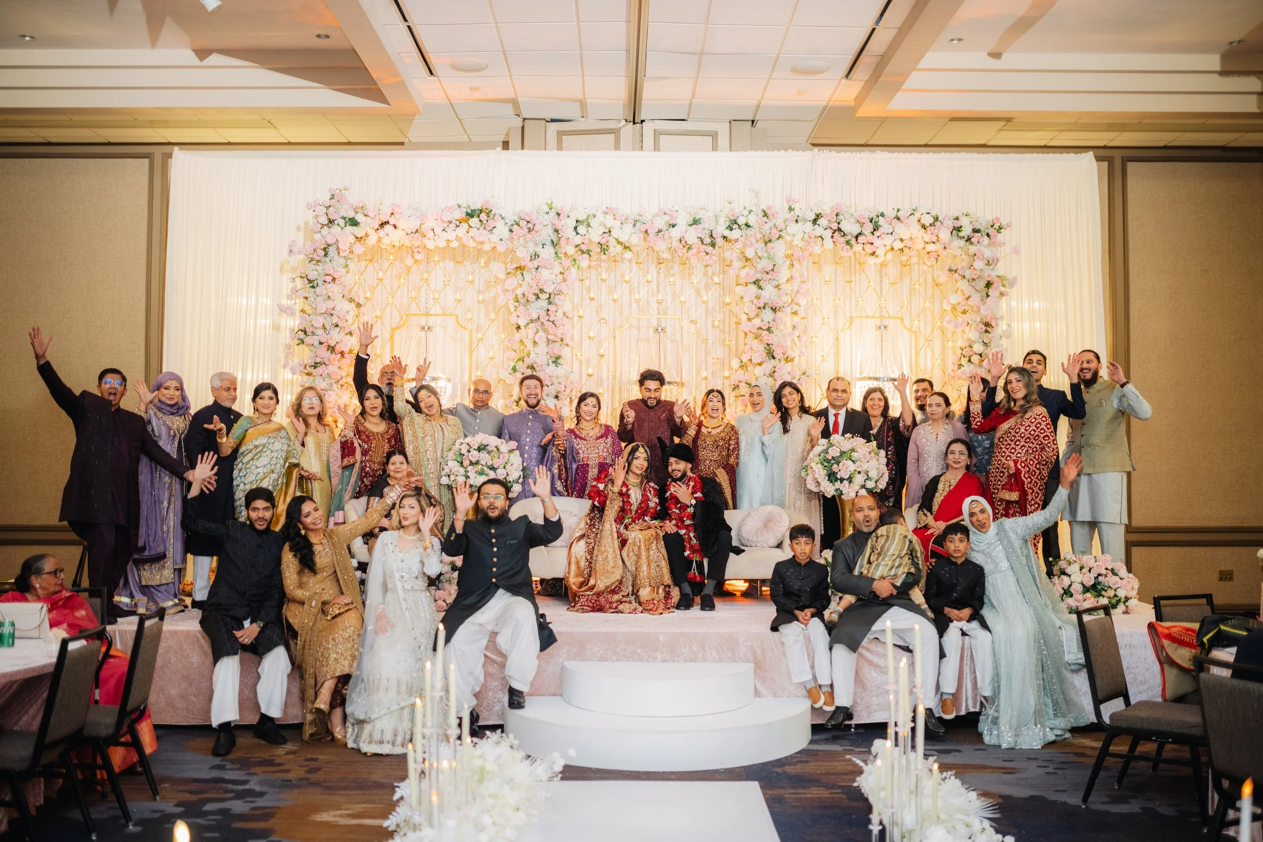A large group of people at a wedding celebration, standing on a stage decorated with pink and white flowers, smiling and posing for the photo in an elegant indoor setting.