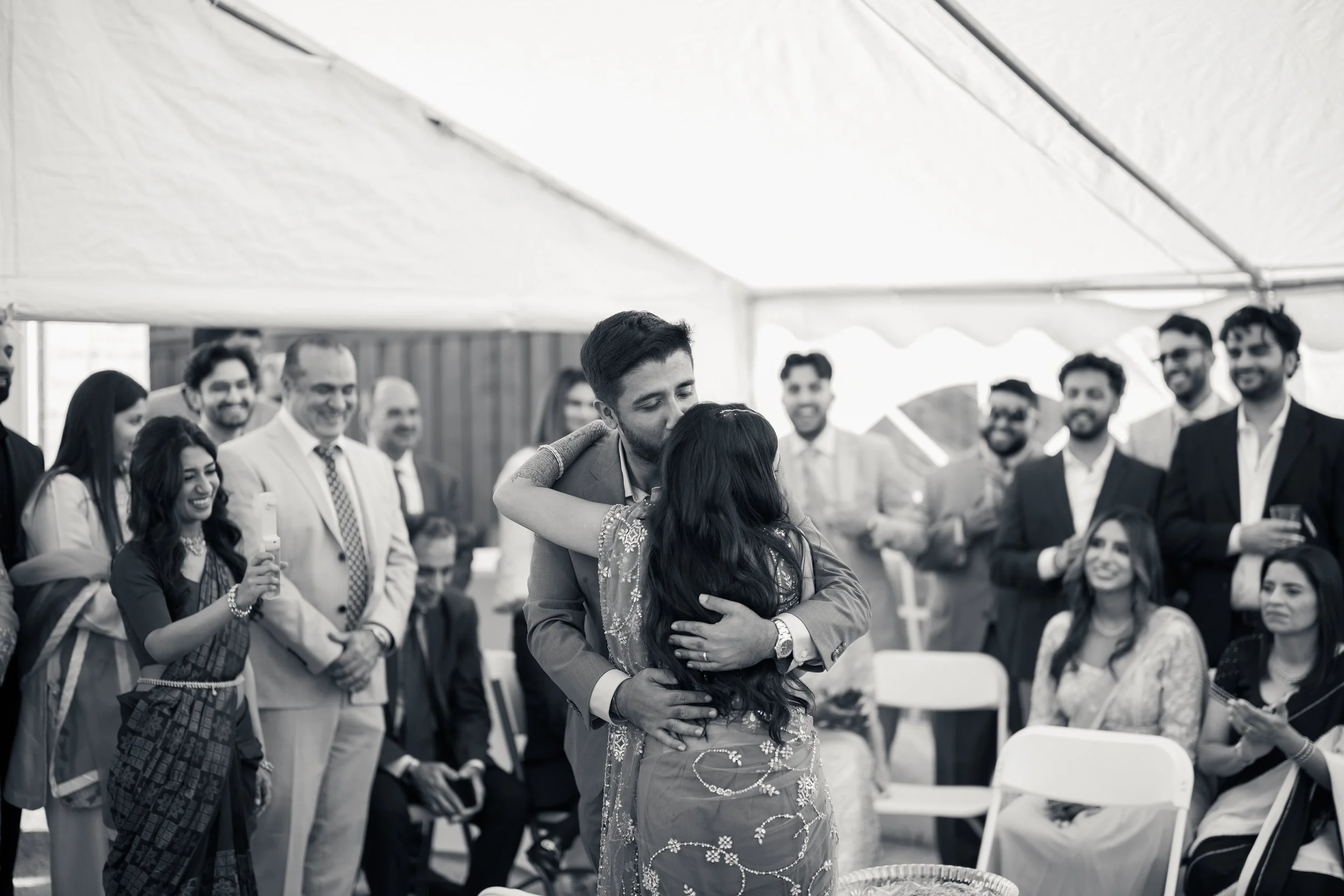 A man and woman hugging at a celebration, surrounded by people dressed in formal attire, under a tent at an outdoor event.