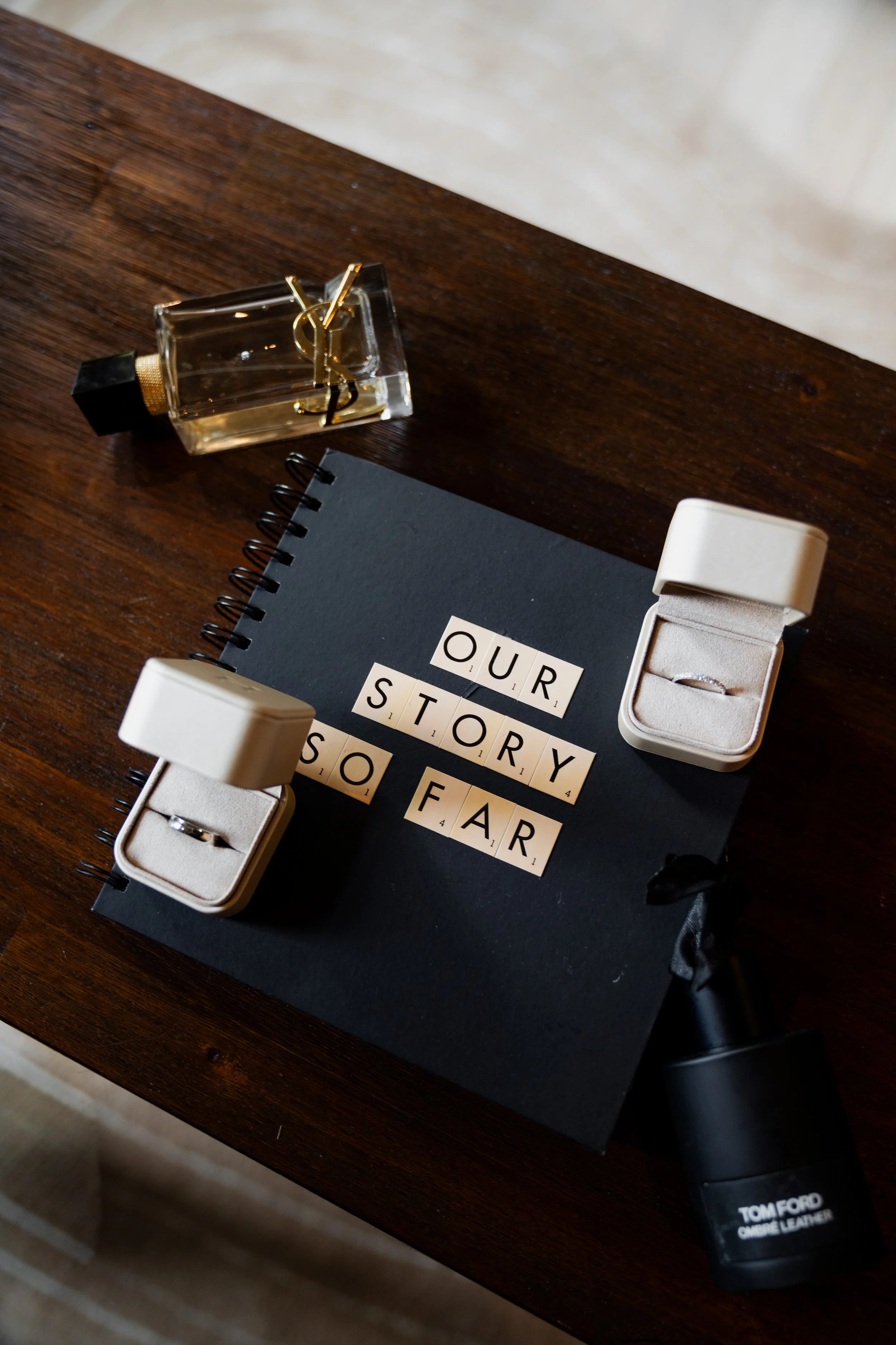 Top view of a wooden table with a perfume bottle, a black notebook with Scrabble-like words, two jewelry boxes containing rings, and a black flashlight labeled "Tom Ford" on a beige surface.